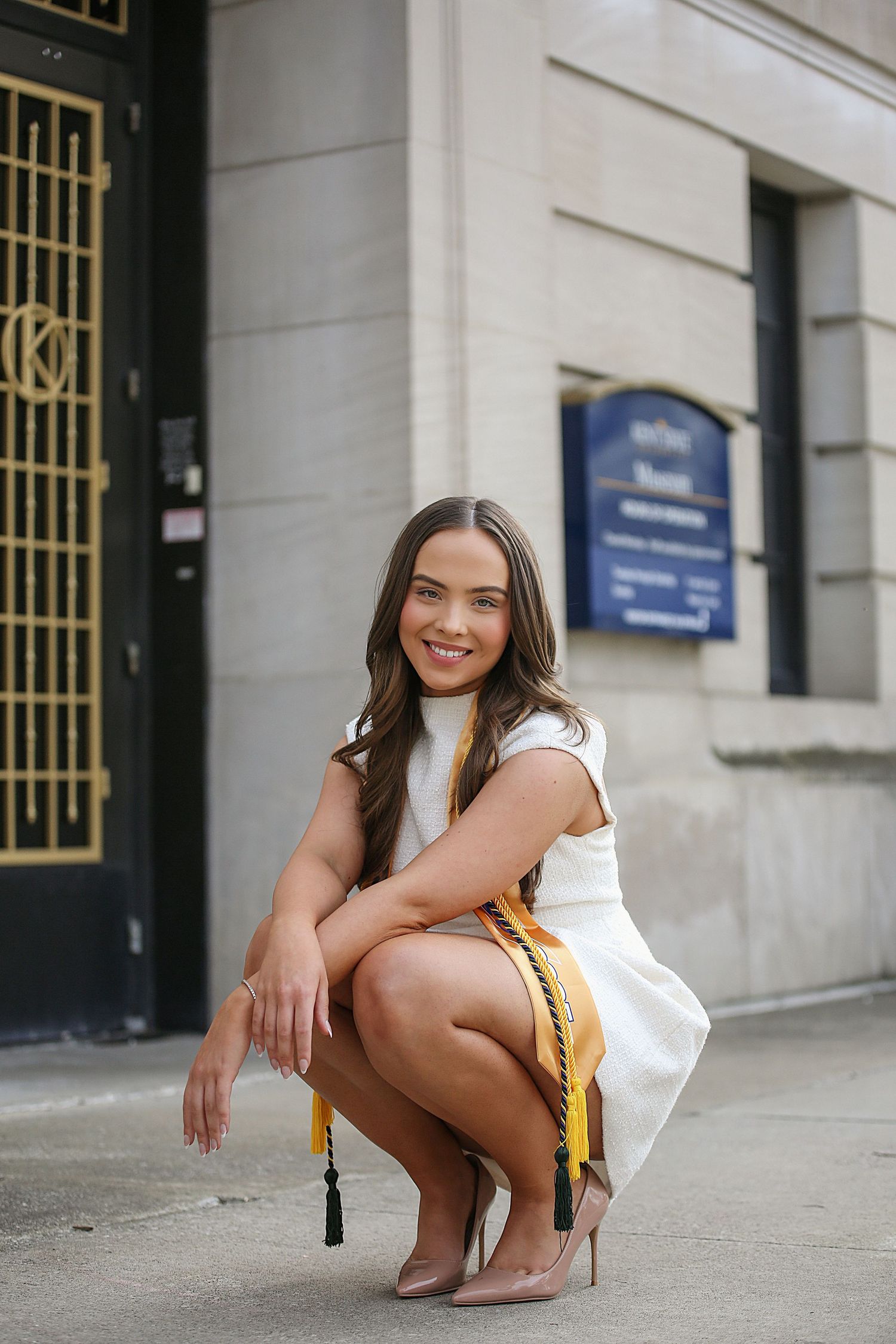 Young woman in white dress crouching on urban sidewalk near decorative gold building details.