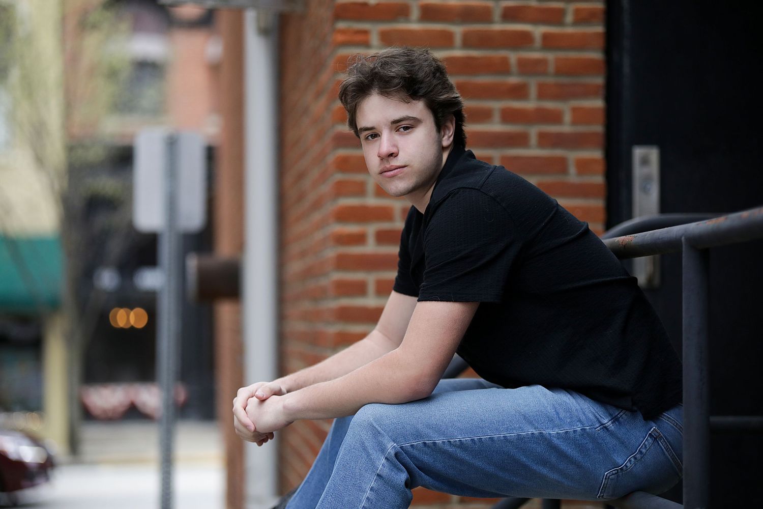 A person in a black shirt and jeans sits casually on an outdoor ledge near a brick wall.