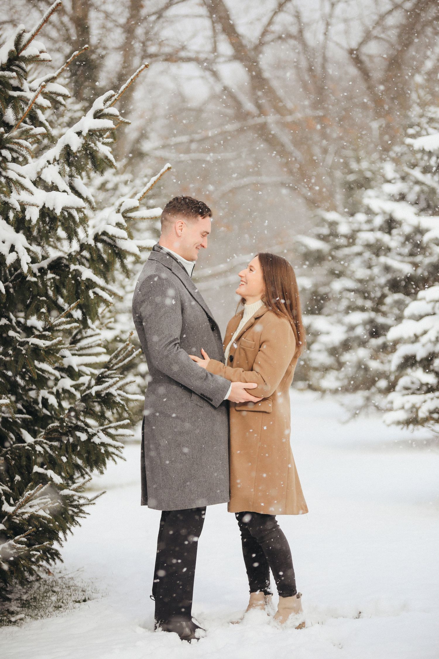 A couple shares a romantic moment in a snowy winter landscape surrounded by snow-covered evergreen trees.