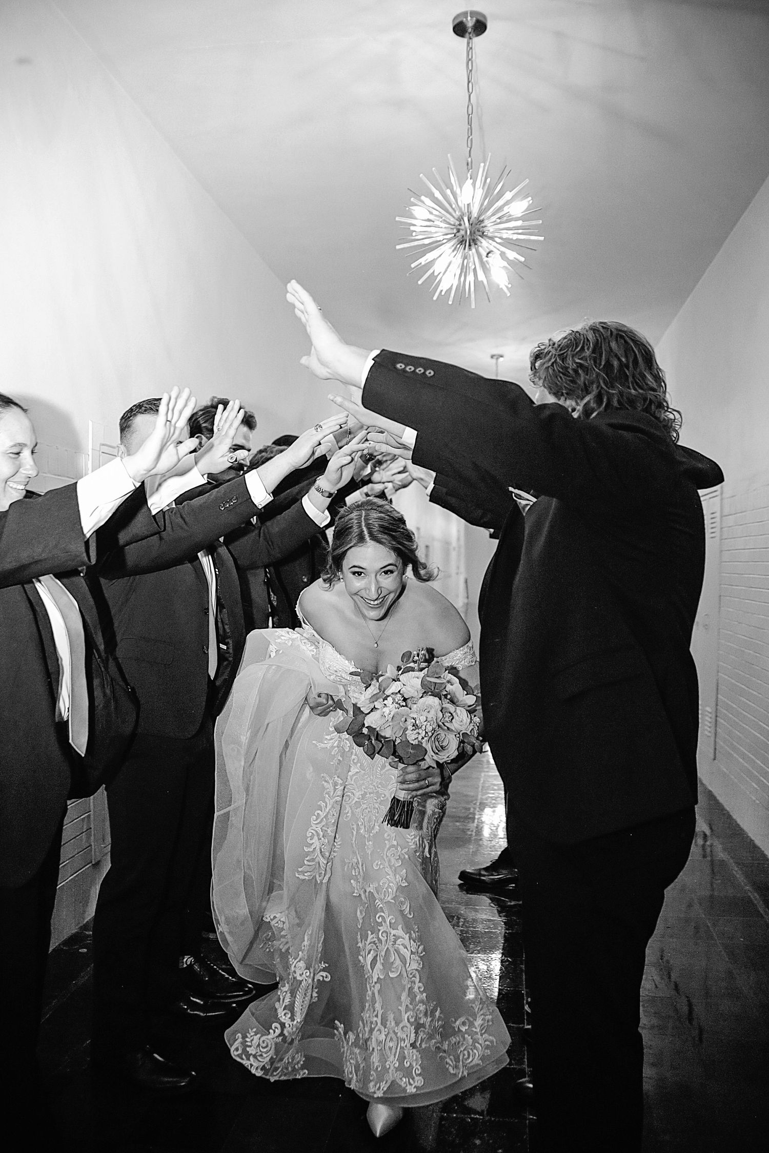 A joyful wedding celebration captured in black and white as guests form a tunnel for the bride walking beneath a modern chandelier.