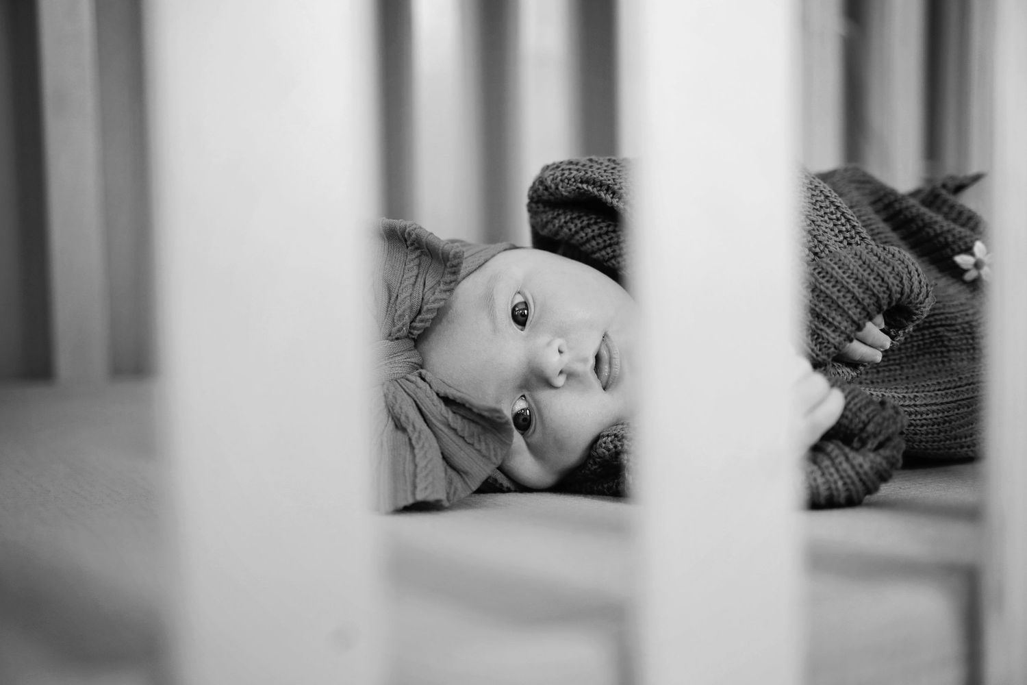 Black and white view through crib bars showing someone resting on a pillow with a cat nearby.