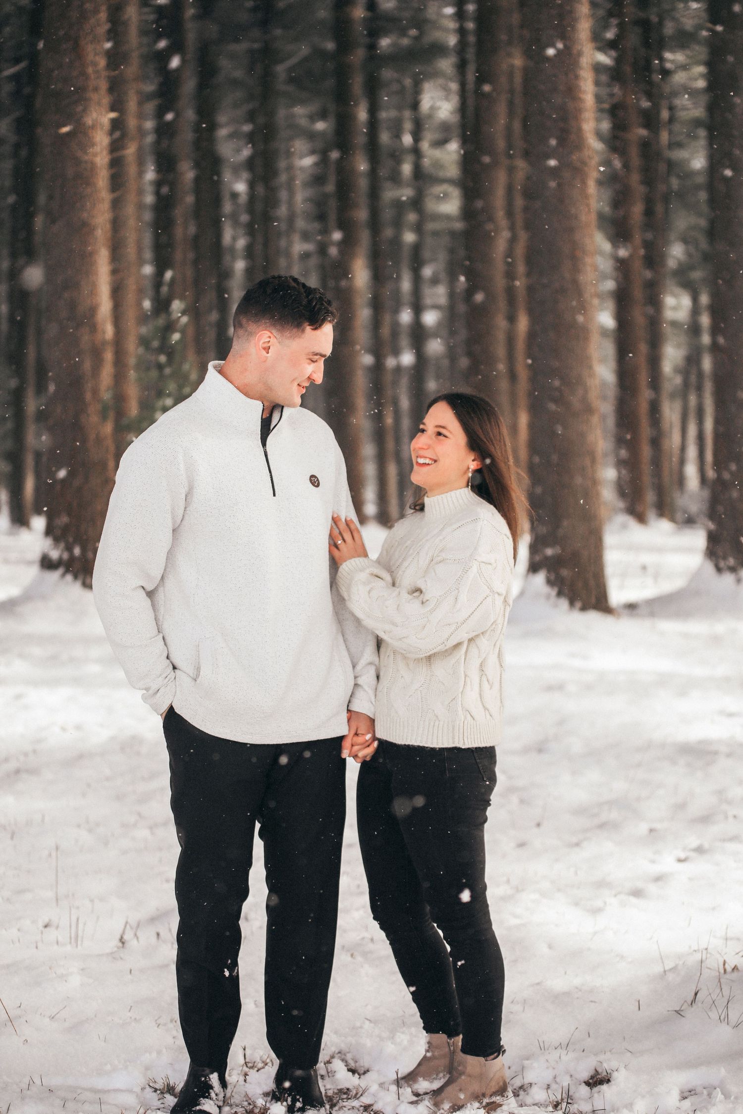 A couple in matching white sweaters and dark pants share a playful moment in a snowy winter forest.