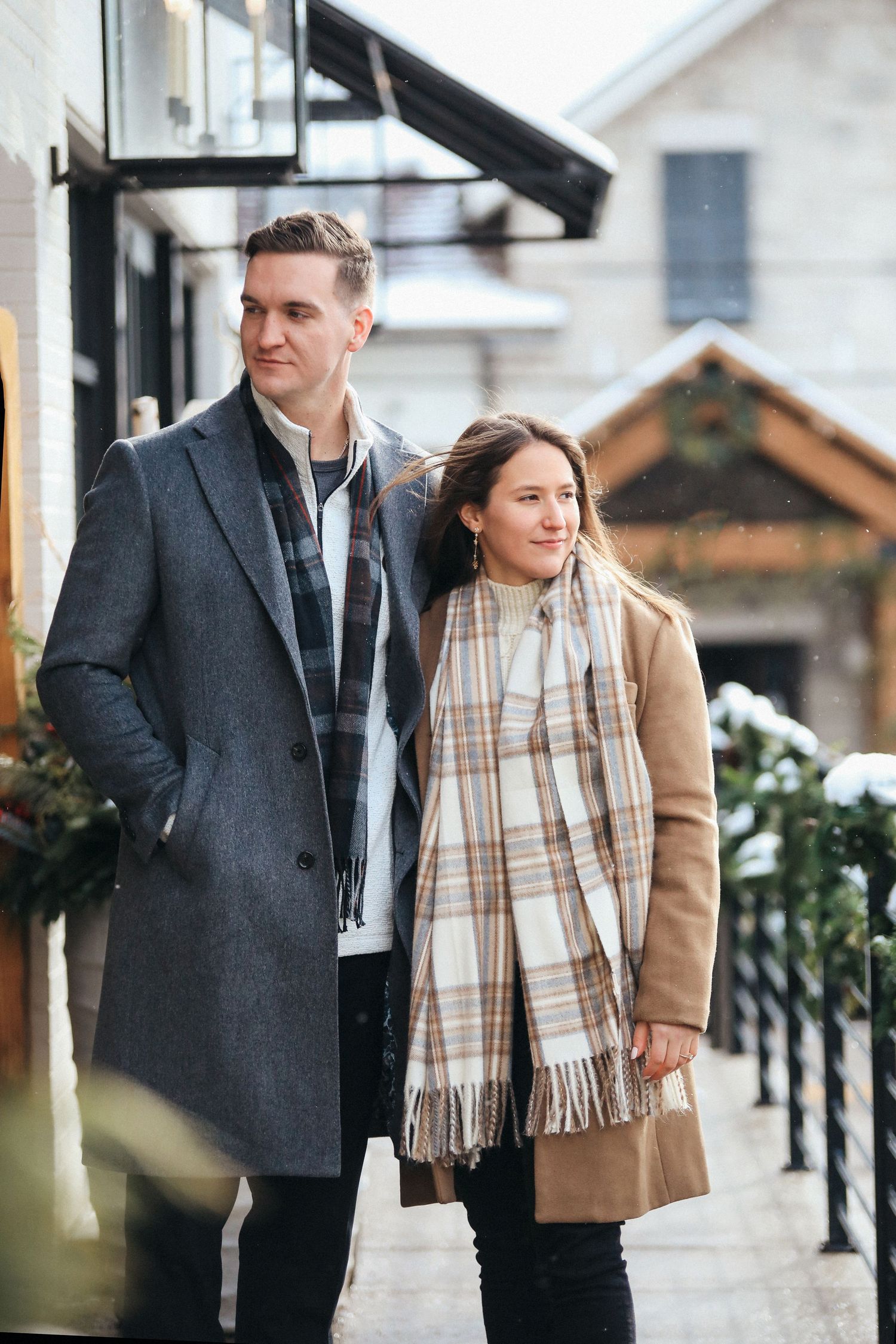 A couple in winter coats strolls together on a sidewalk, wearing stylish outerwear and plaid scarf on a cool day.