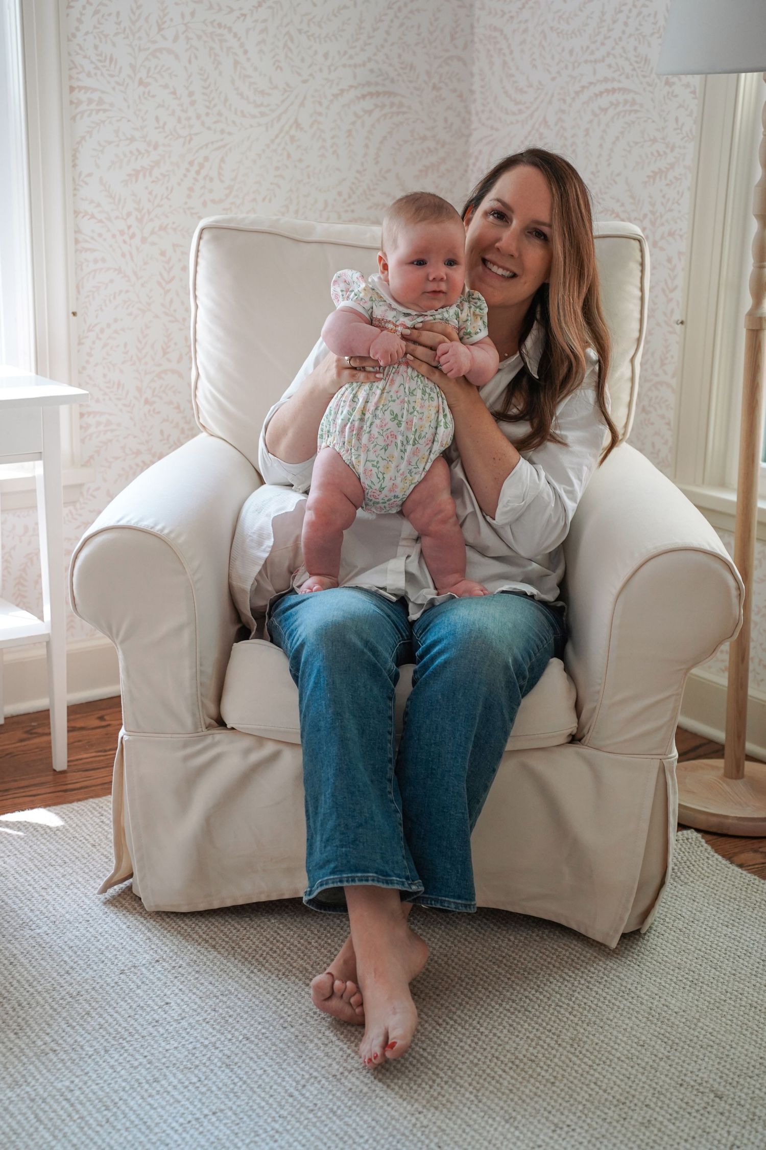 A parent and baby dressed in matching light colors sit together in a cozy white armchair in a bright nursery setting.