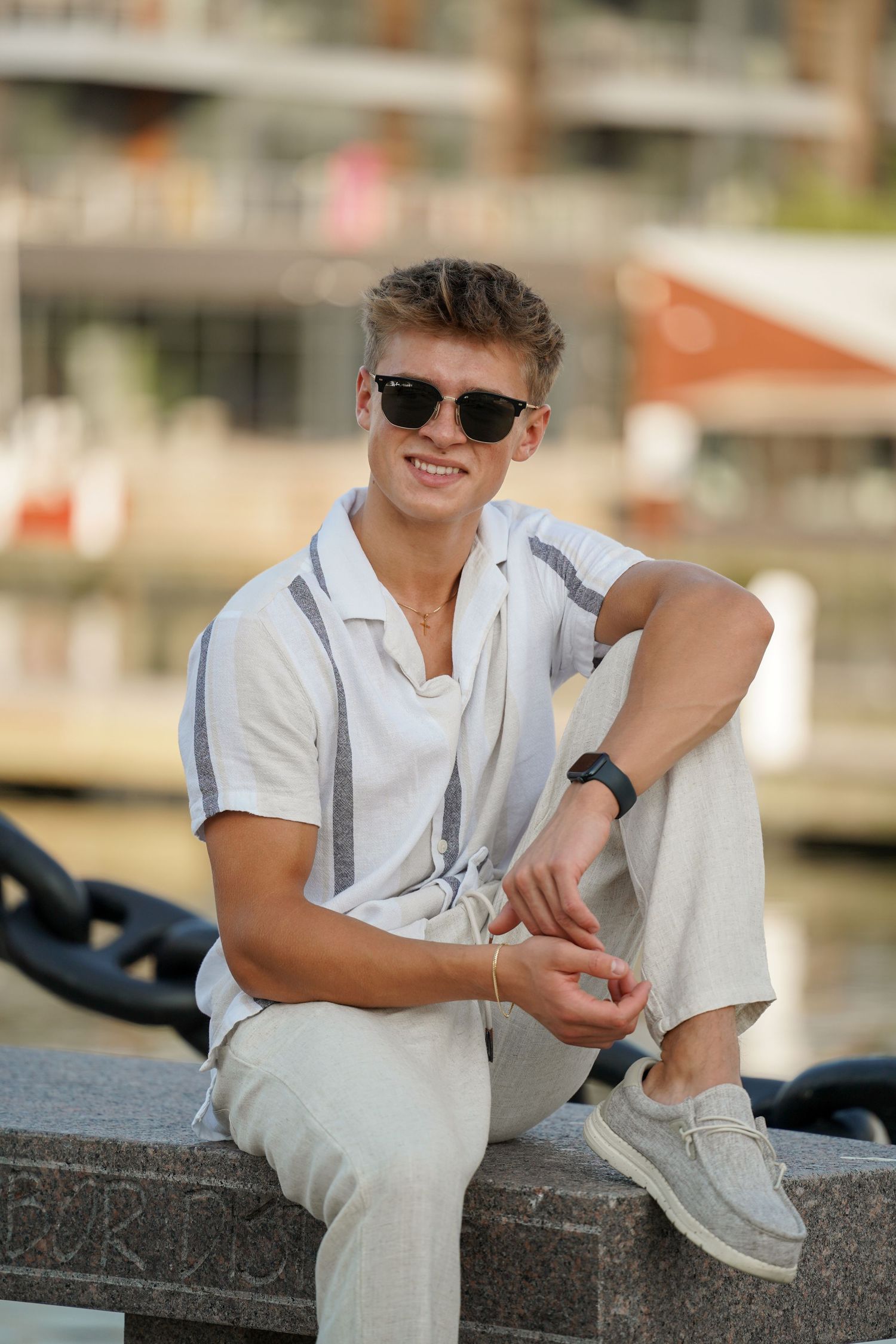 A person wearing sunglasses and a white polo shirt sits casually on a stone ledge outdoors on a sunny day.