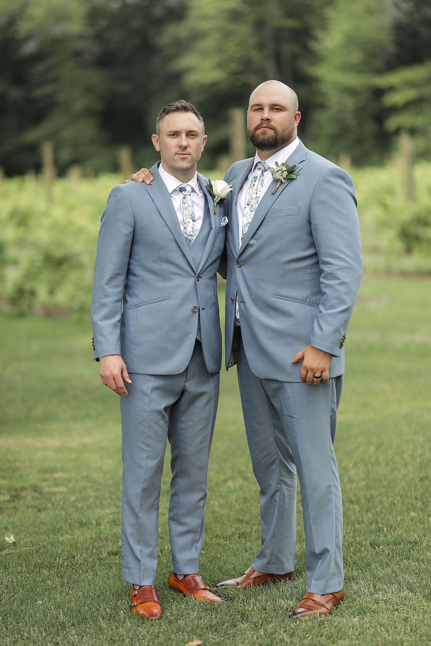 Two groomsmen pose together wearing matching light gray suits and brown shoes in an outdoor garden setting.
