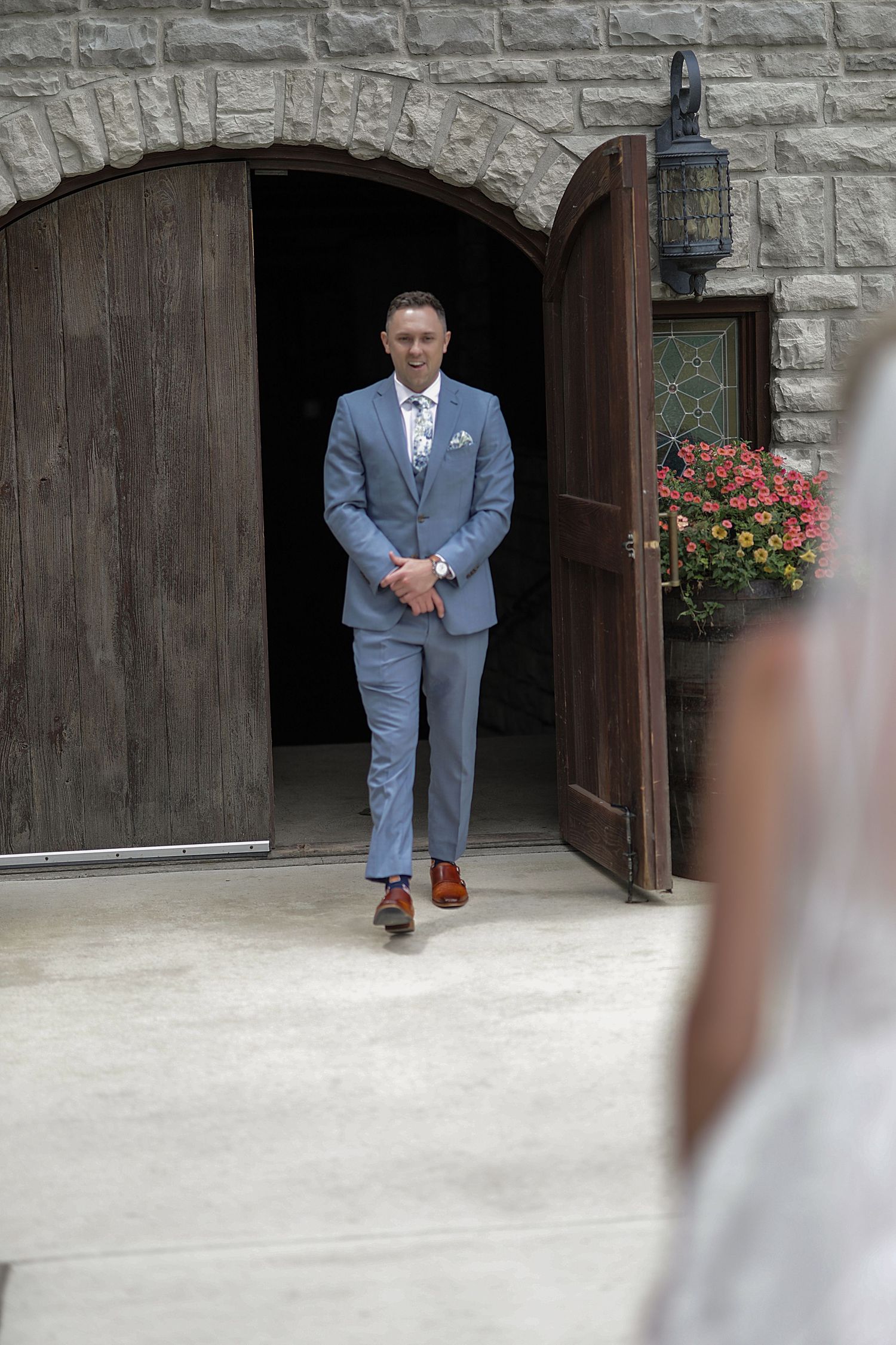 A suited figure stands in a stone archway doorway wearing a light blue suit and boutonniere.