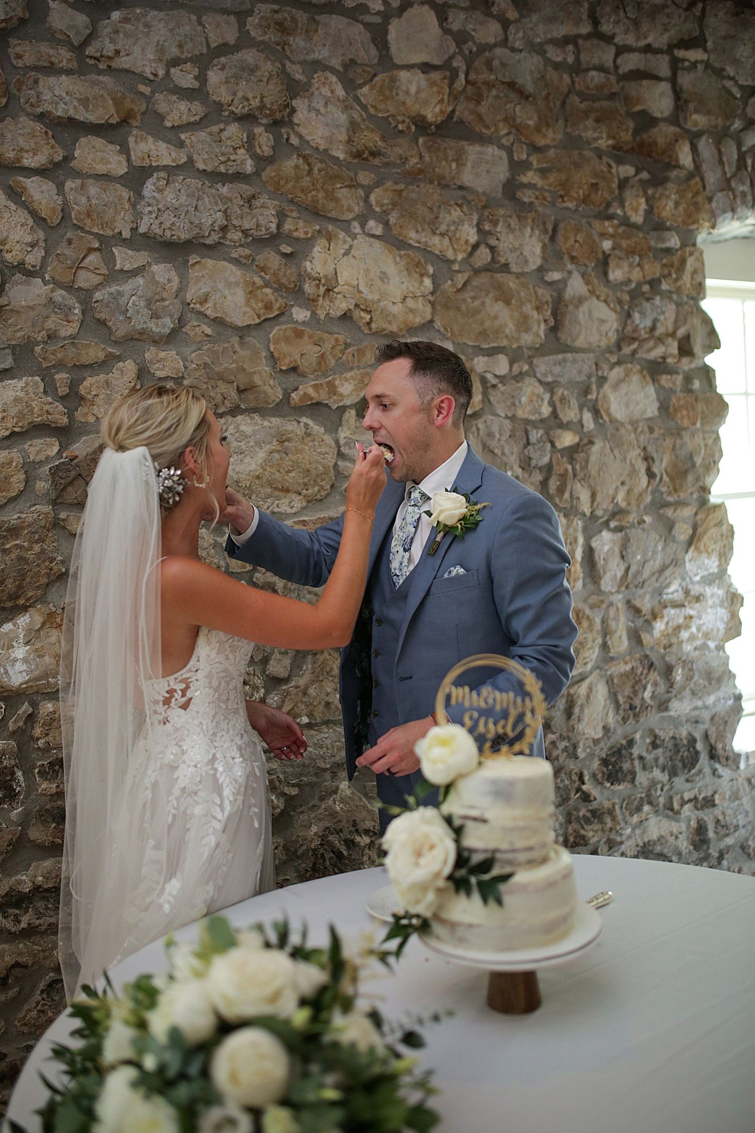 Newlyweds share cake-feeding moment against rustic stone wall backdrop.