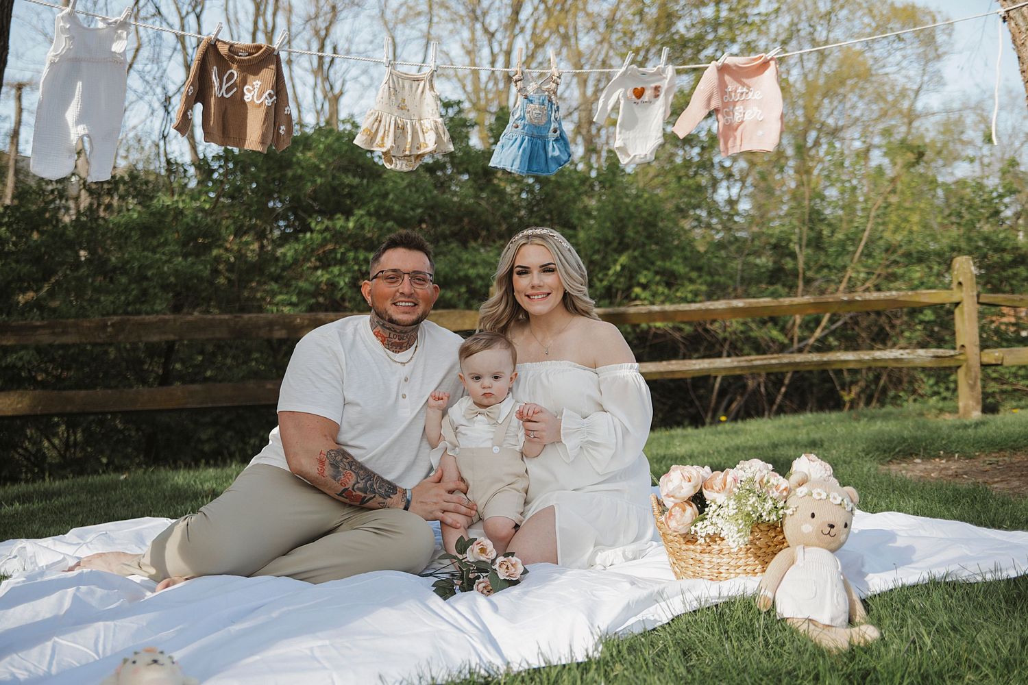 A family enjoys a picnic on a white blanket with laundry hanging on a clothesline in the background.