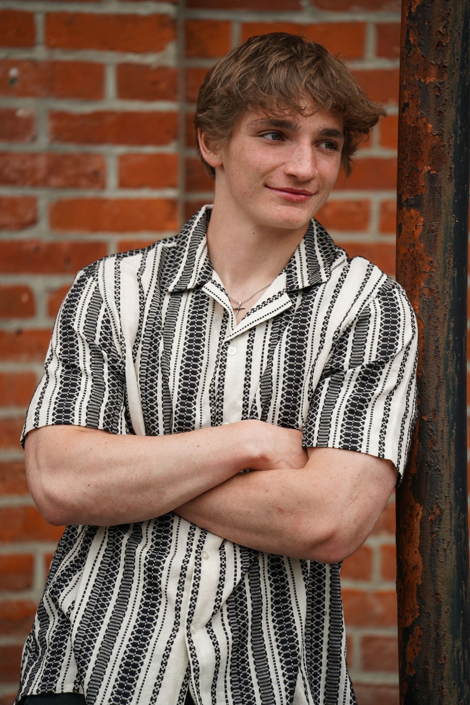 A person in a black and white striped button-up shirt poses with crossed arms against a brick wall background.