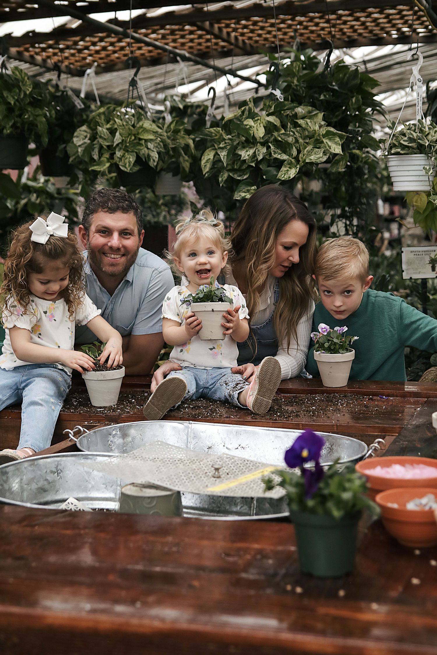 Family enjoys planting activities together at a rustic wooden table in a greenhouse with potted plants and gardening supplies.