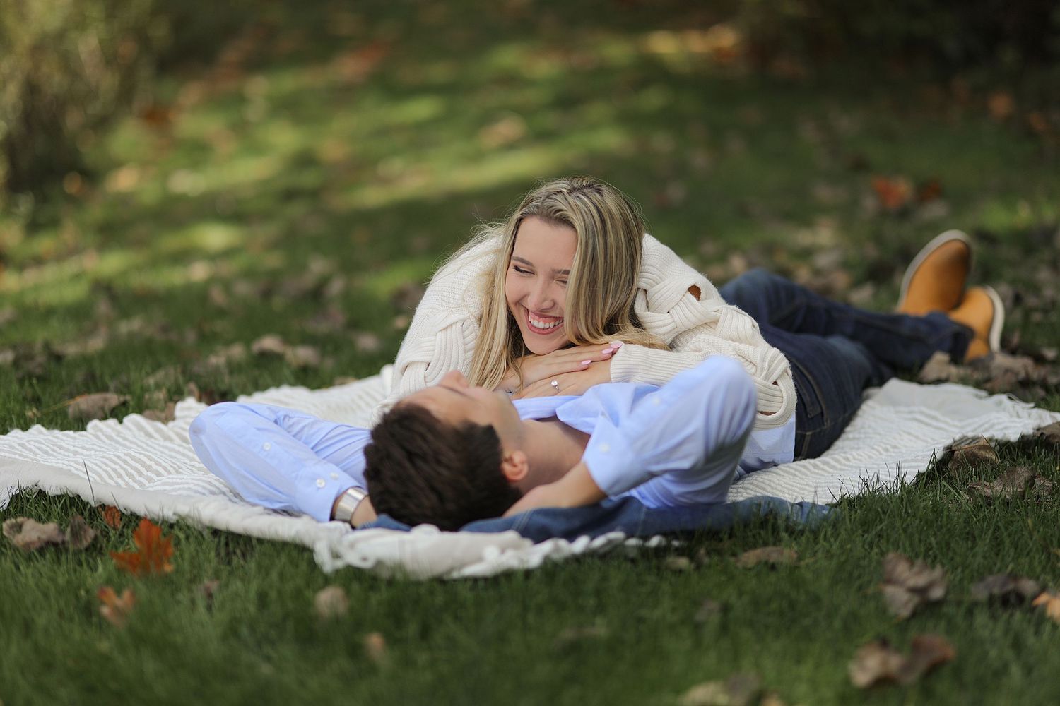 A couple laughs together while laying on a blanket in dappled sunlight on grass during an outdoor photo session.