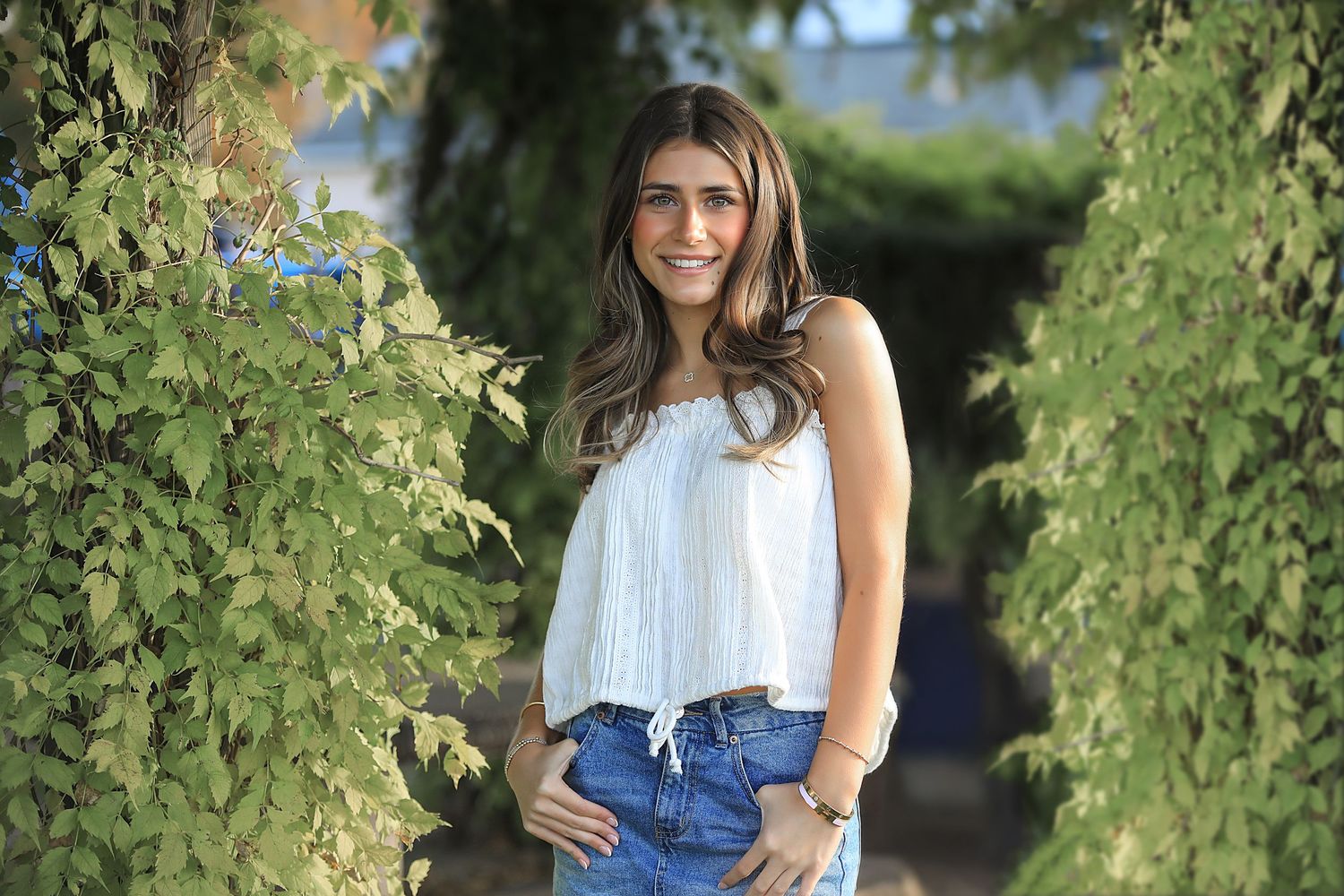 A person in a white off-shoulder top and denim shorts poses between green foliage on a sunny summer day.