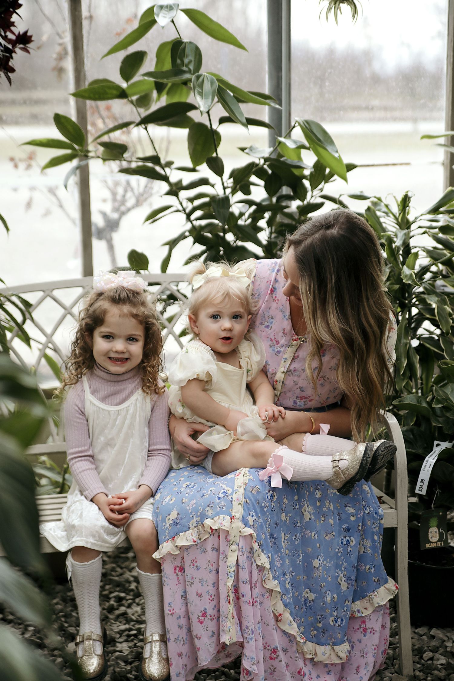 Family wearing floral dresses shares tender moments together by large windows surrounded by indoor plants.