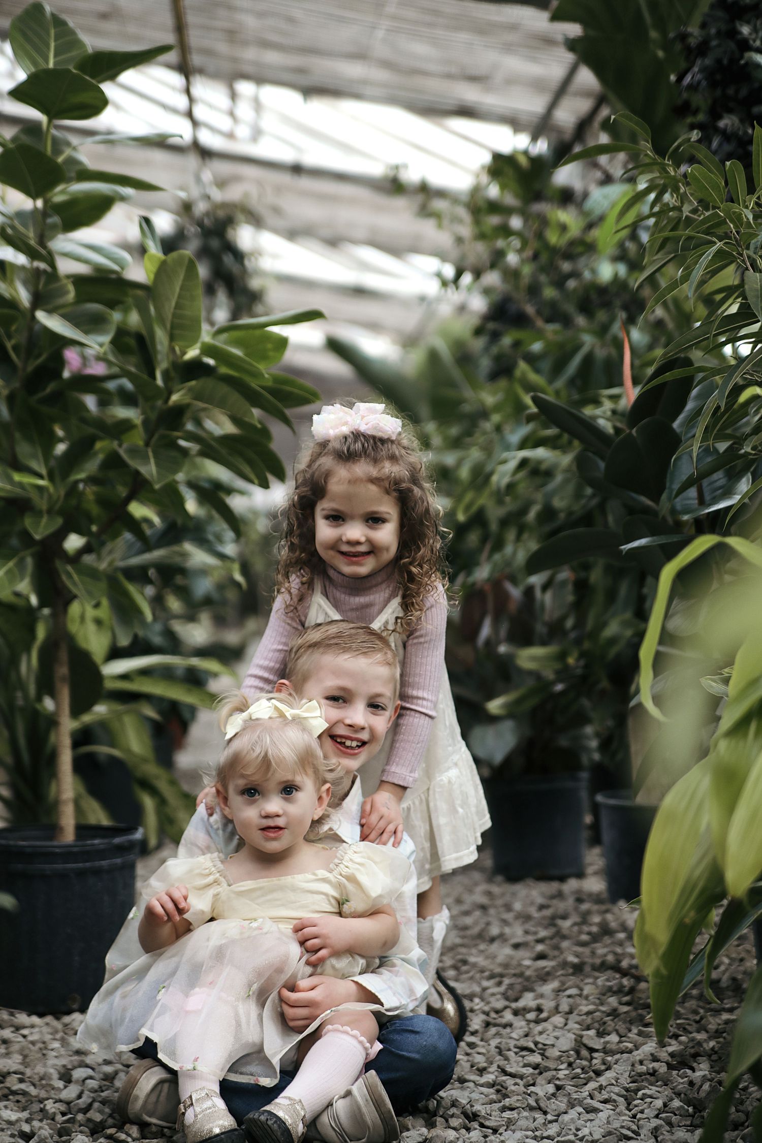 Children sitting together among potted plants in a greenhouse with natural light streaming through the ceiling.