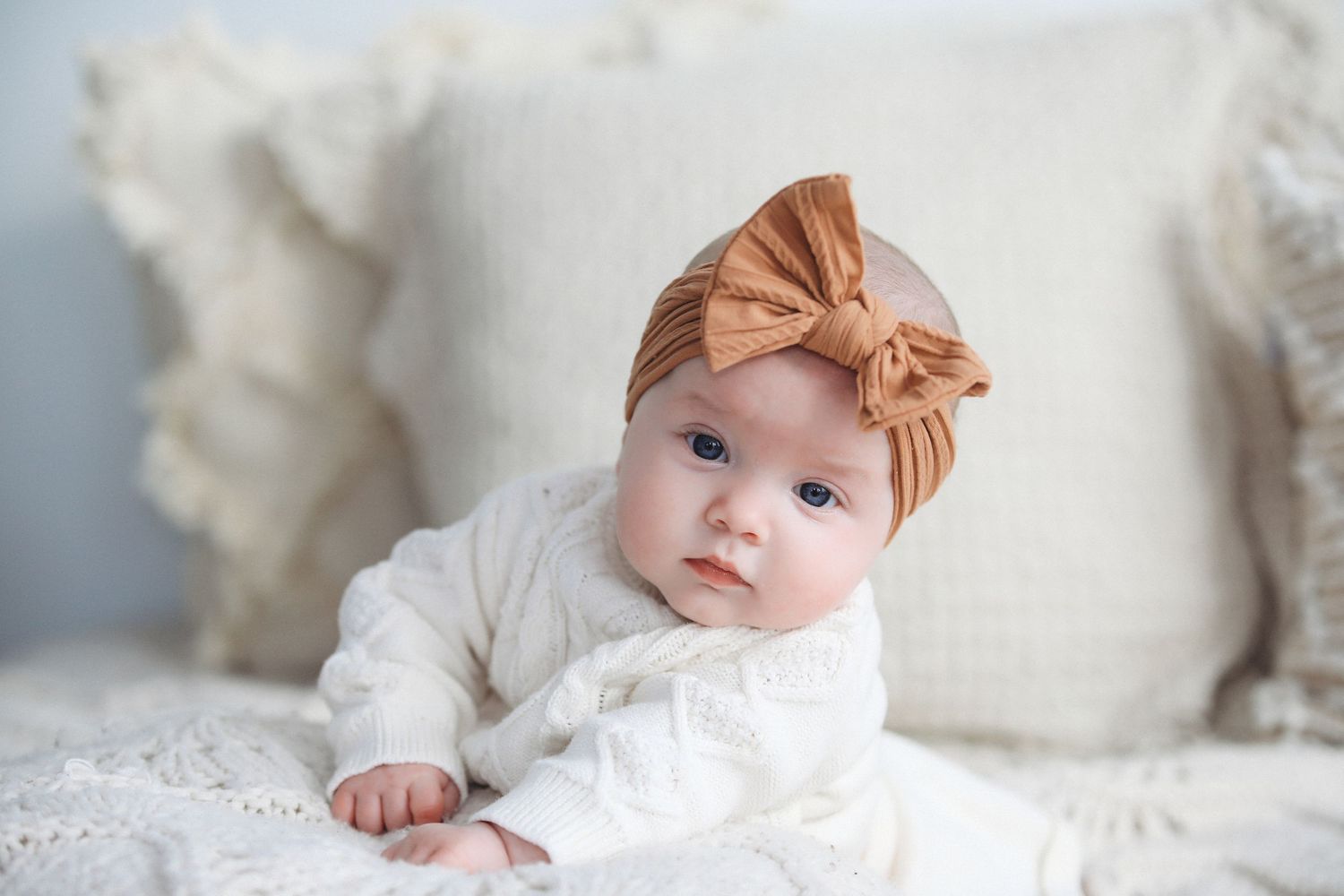 Baby wearing a white dress and brown bow headband lying on white bedding.