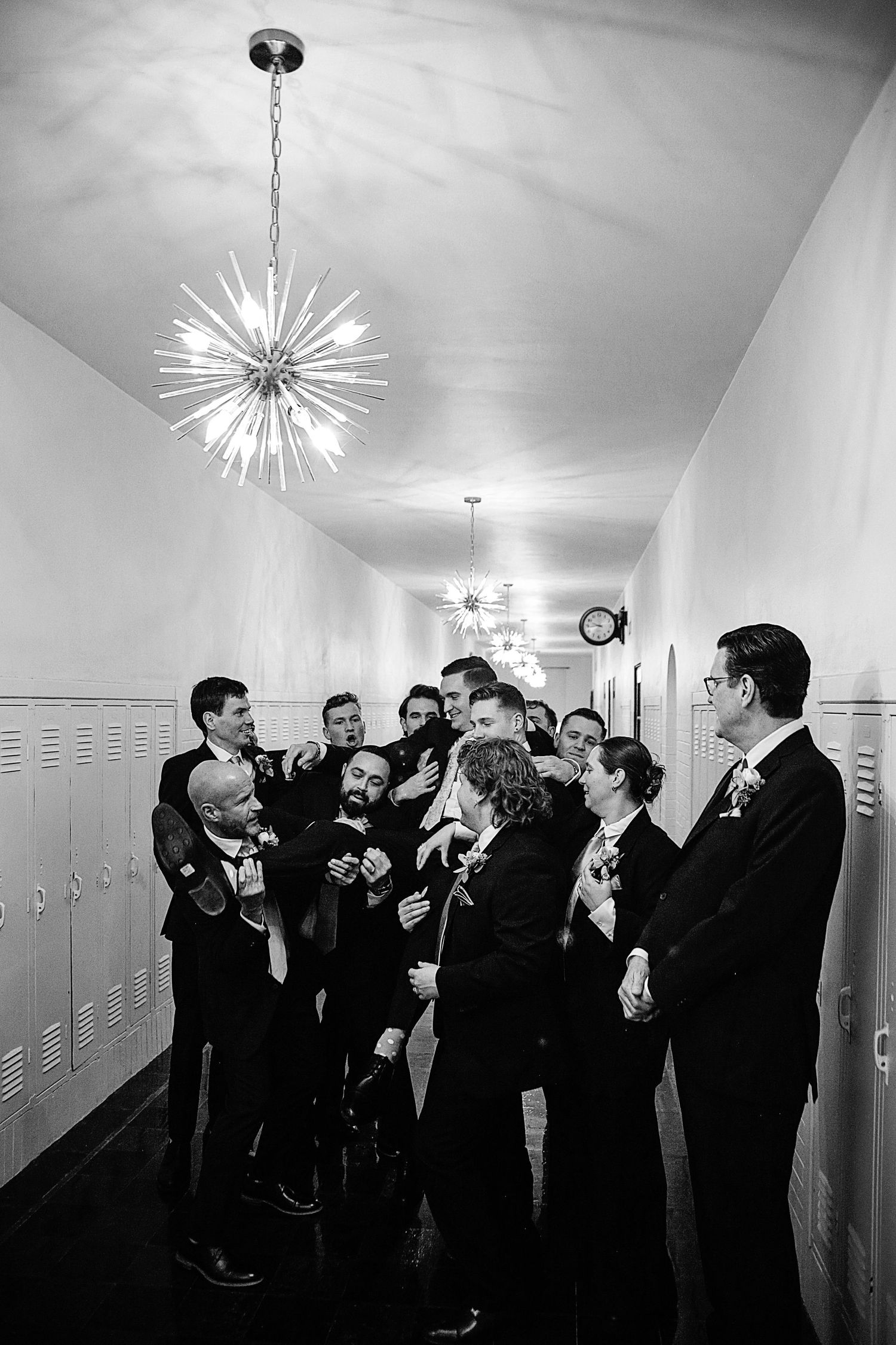 A group of groomsmen in black tuxedos gather in a hallway under modern light fixtures.