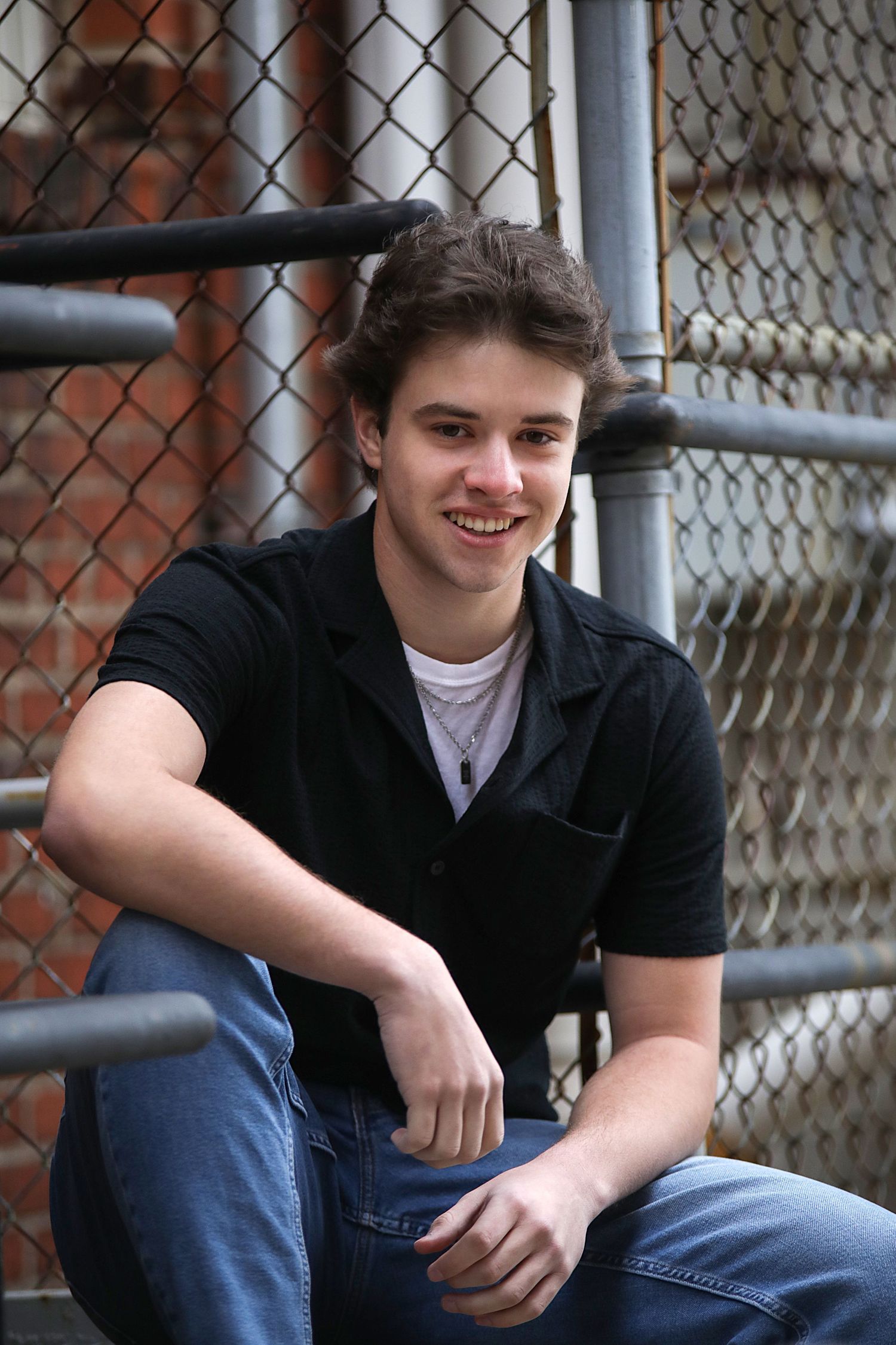 A person in casual attire sits against a chain link fence wearing blue jeans and a black shirt.