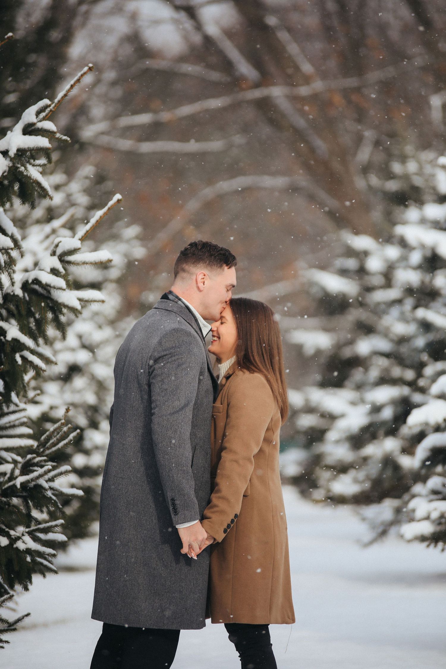 A couple shares an intimate moment in a snowy winter forest setting while wearing warm coats.