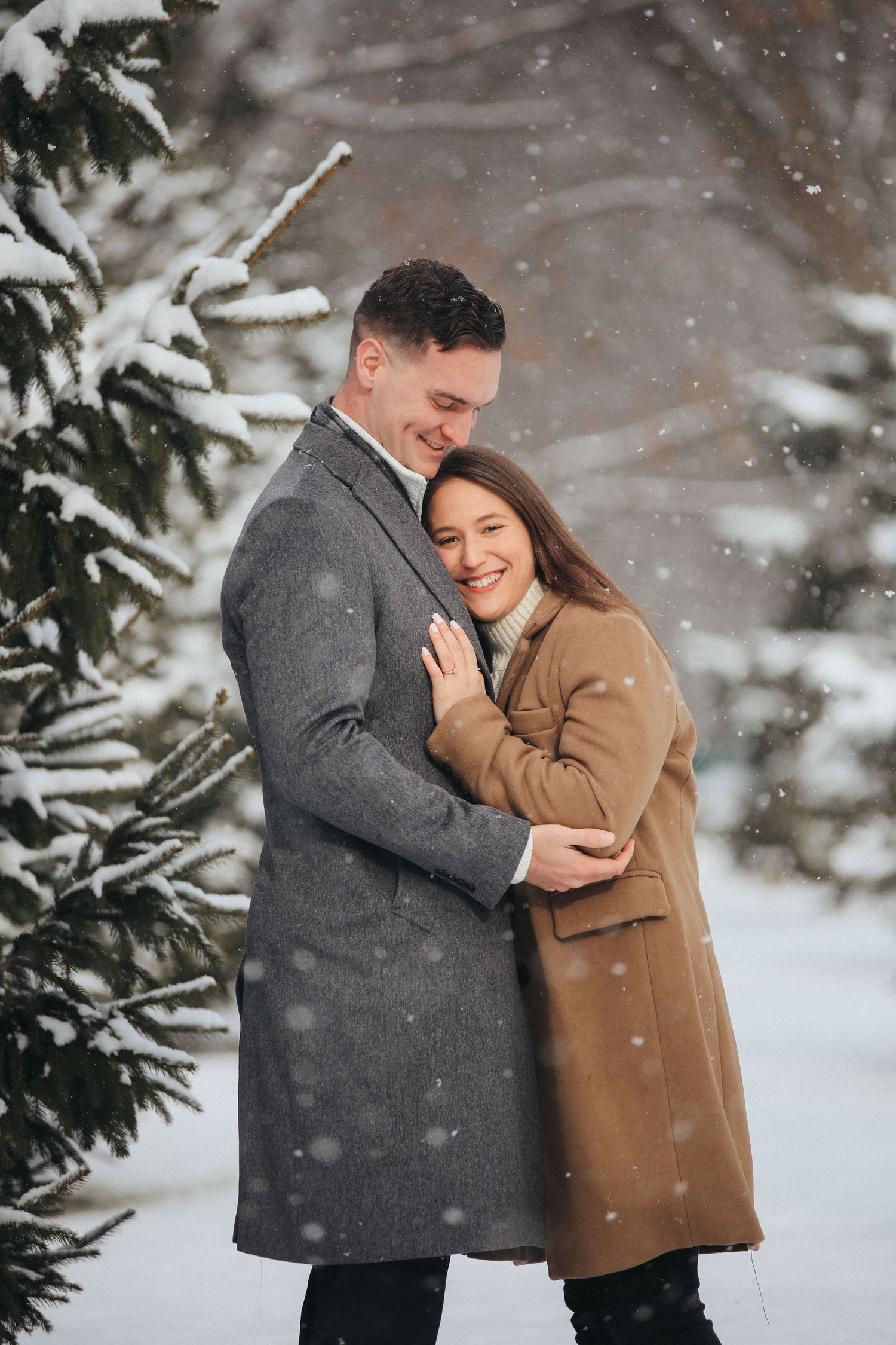 A couple shares an intimate embrace in a snowy winter setting with evergreen trees in the background.