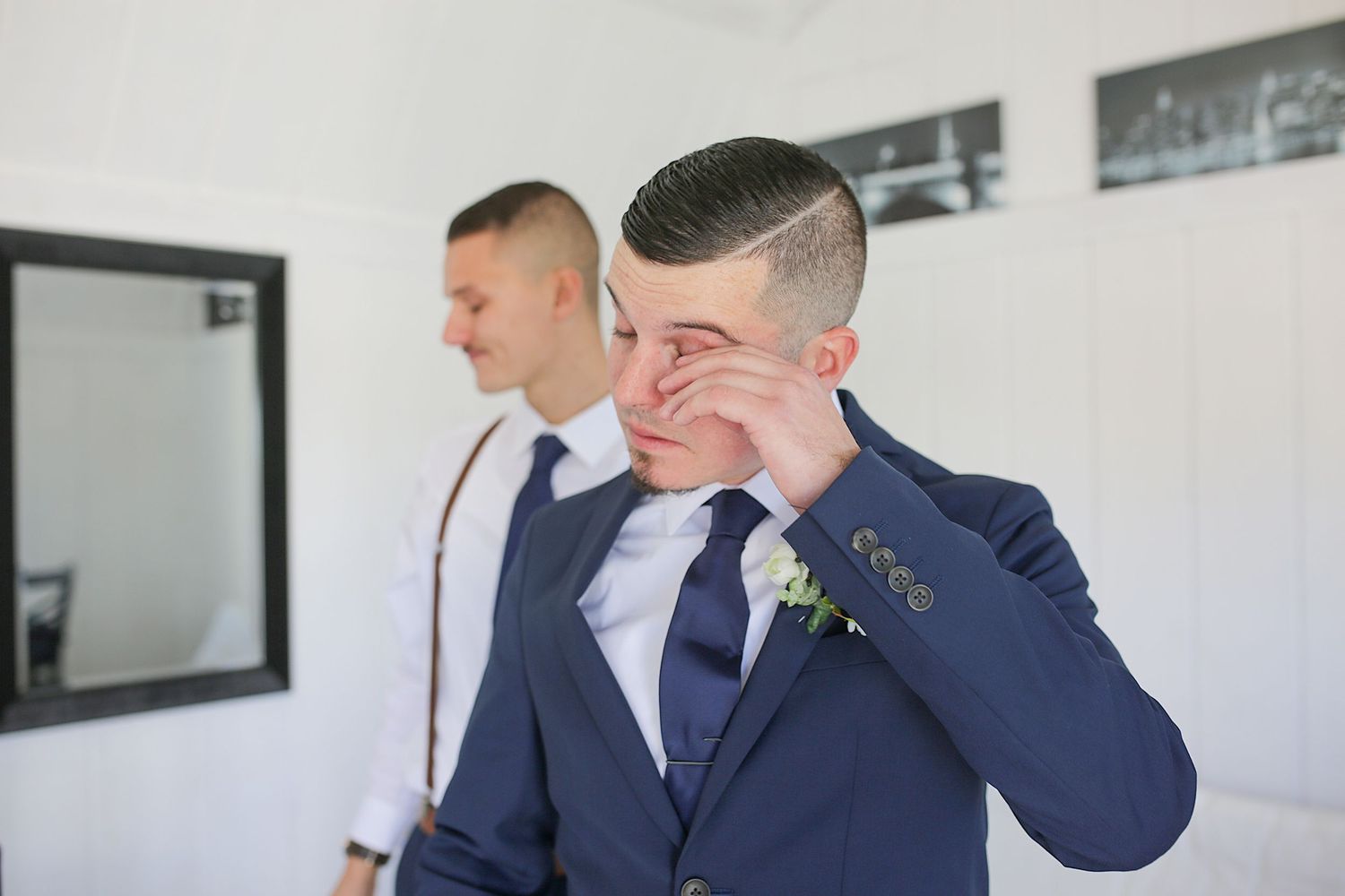 Emotional moment caught in navy suit while someone stands in background against white wall with mirror.