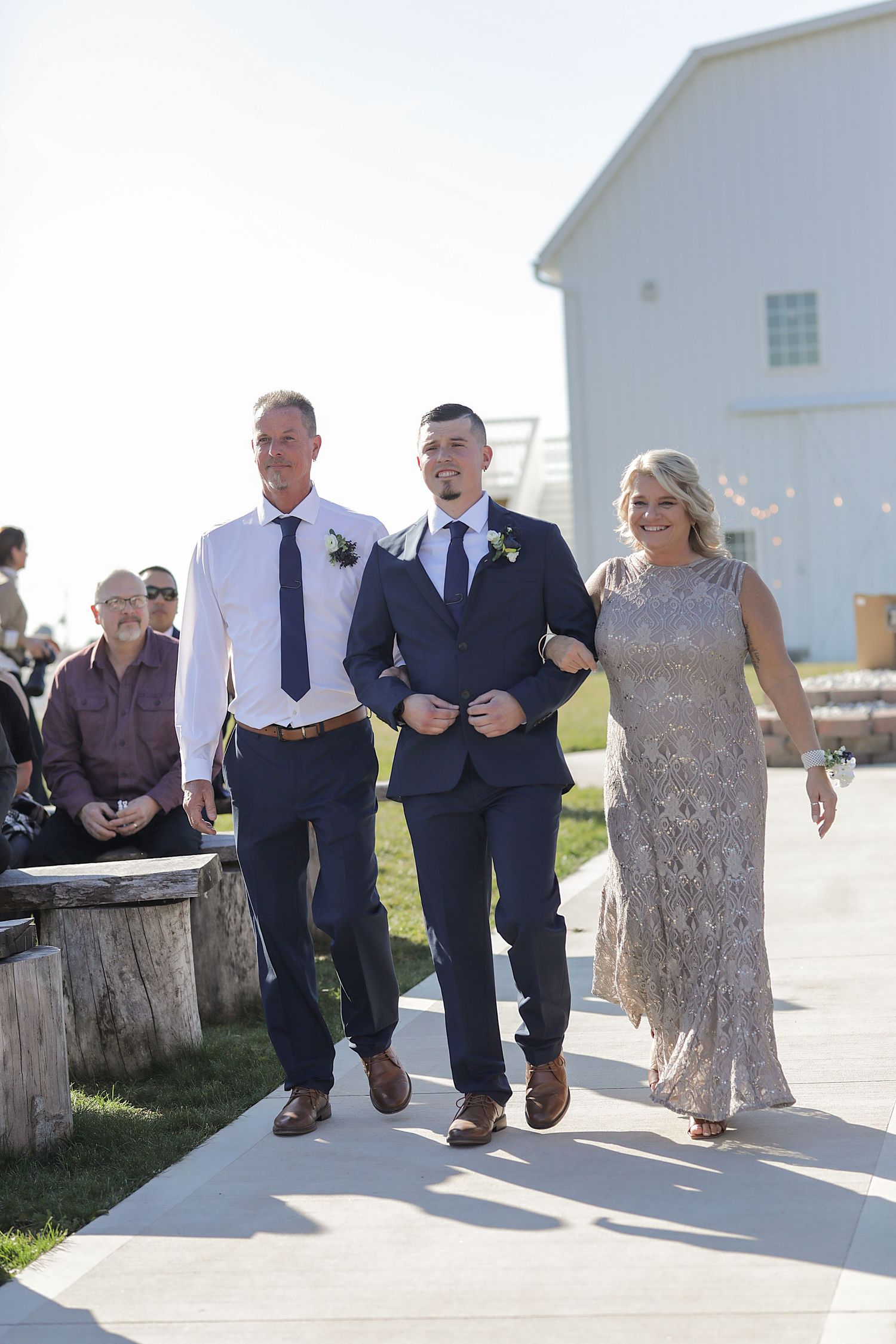 Wedding party members walk down a path beside a white barn on a sunny day.