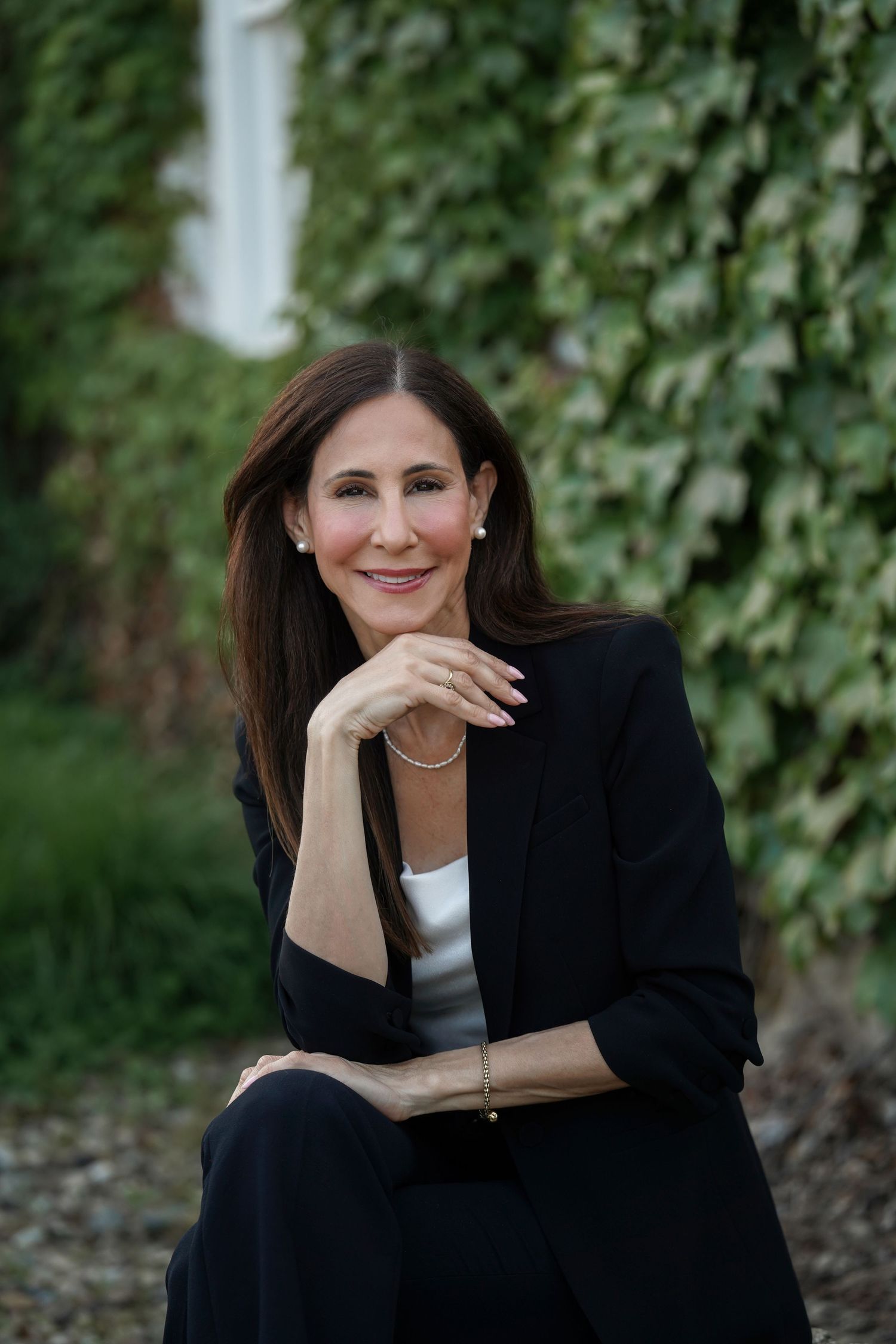 Professional portrait of someone in a black blazer and white top seated outdoors with ivy-covered wall in background.