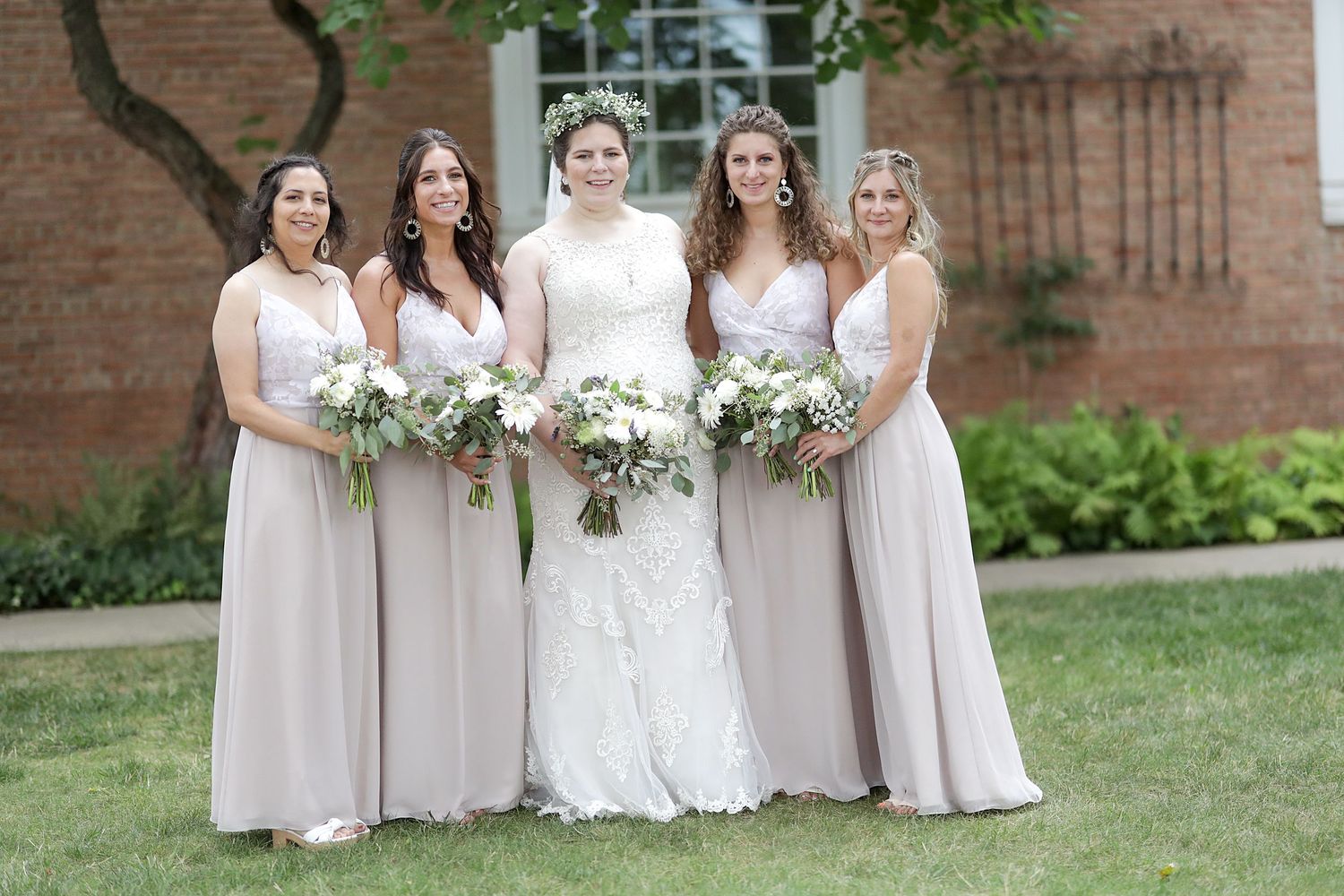 A bridal party in blush pink dresses and white gowns poses together outdoors near a brick building.