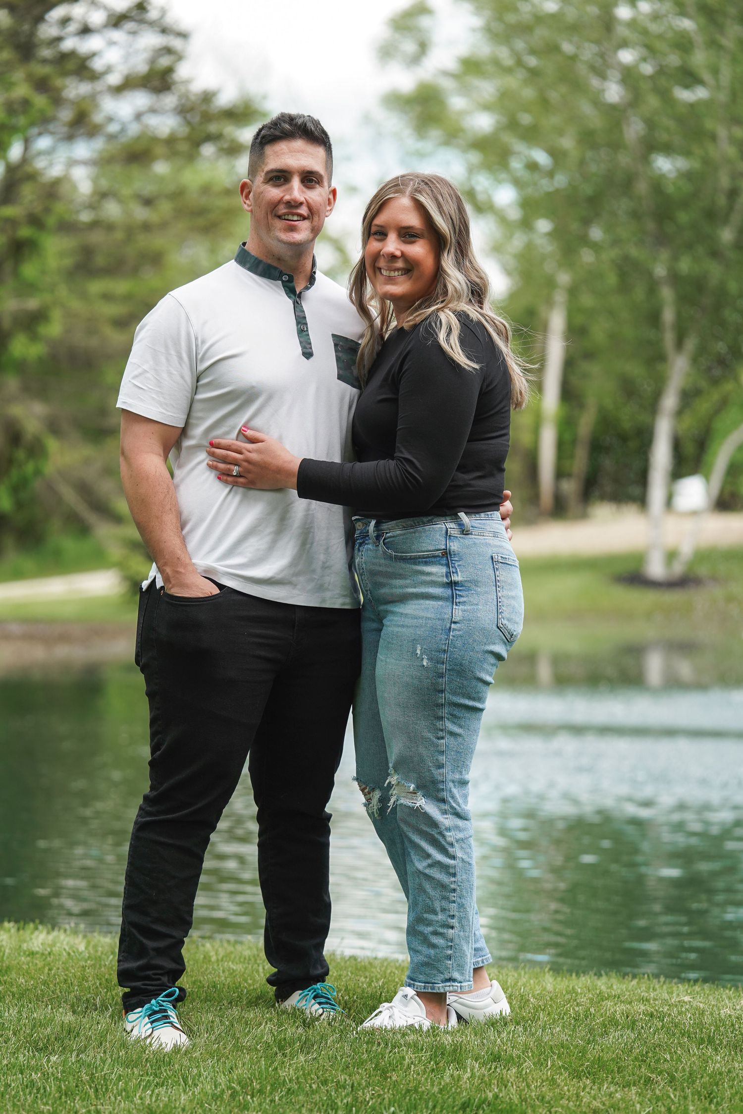 A couple stands together outdoors near a serene lake, enjoying a tender moment on a lush green lawn.