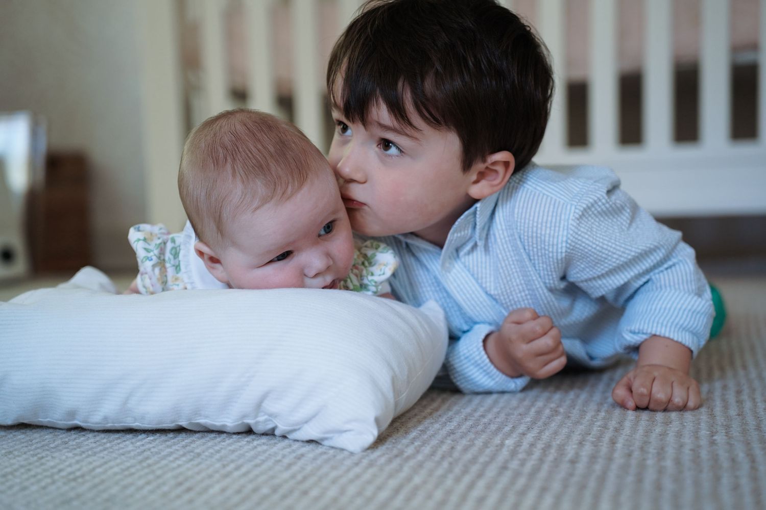 Two small children lying on a carpet play together with toys and interact in a warmly lit room.