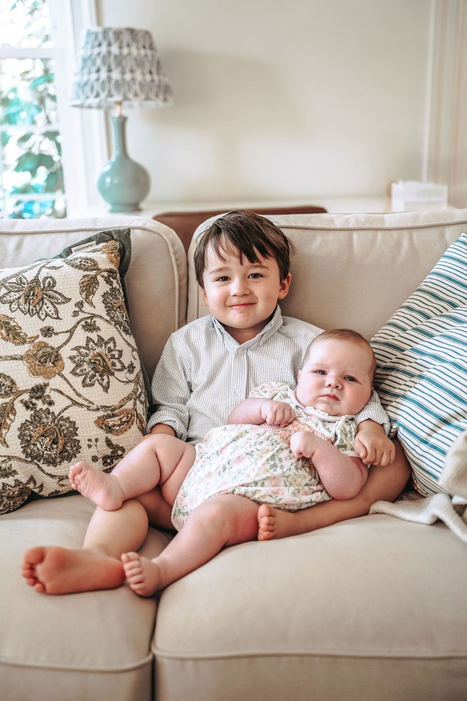 Two children sit together on a beige couch with decorative throw pillows and a table lamp in the background.