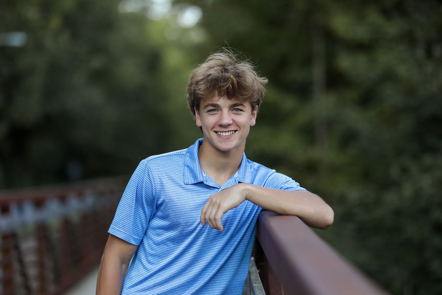 Young person in light blue polo shirt smiling while leaning on outdoor railing with greenery in background.