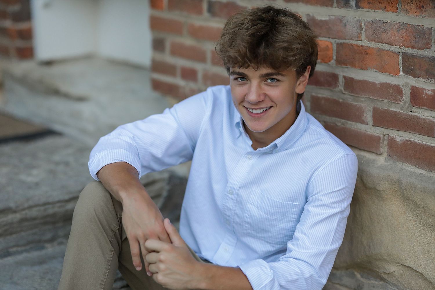 Young person in white button down shirt sits casually against brick wall while smiling outdoors.