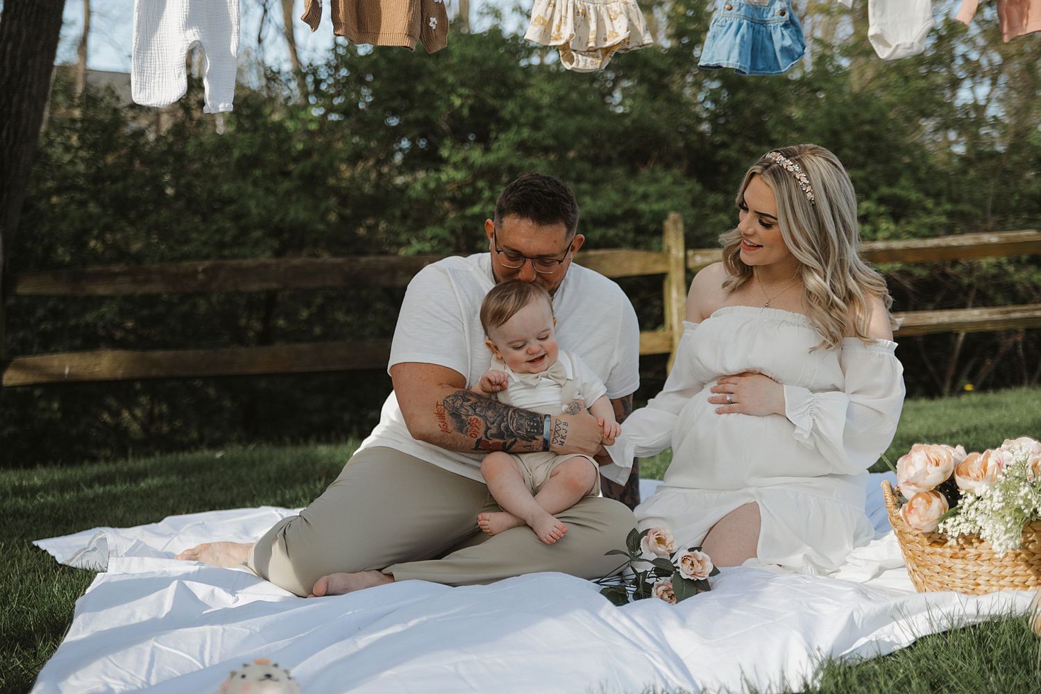 Expectant parents sitting on a white blanket in a garden setting with flowers and laundry hanging in the background.