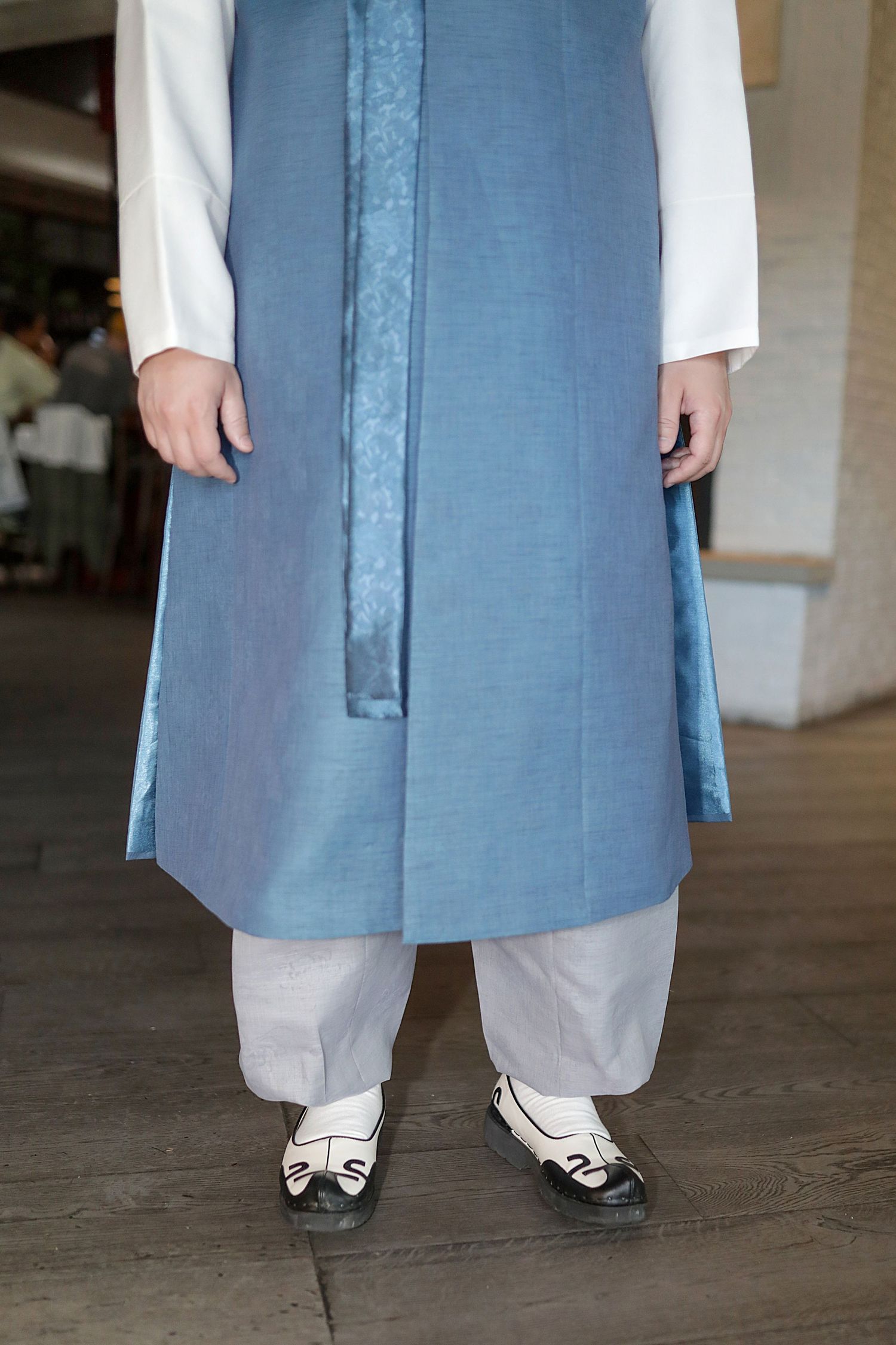 A person wearing a light blue traditional kurta with white pants and black and white shoes stands on a wooden floor.