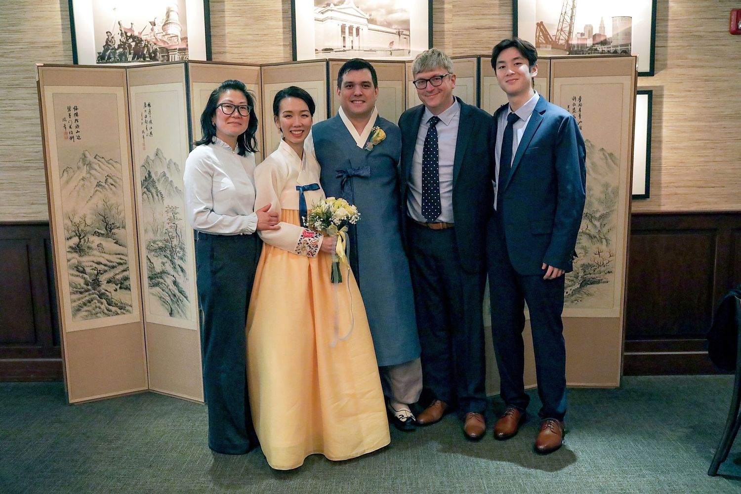 A group of people stand together in formal attire against a backdrop of decorative folding screens with Asian artwork.