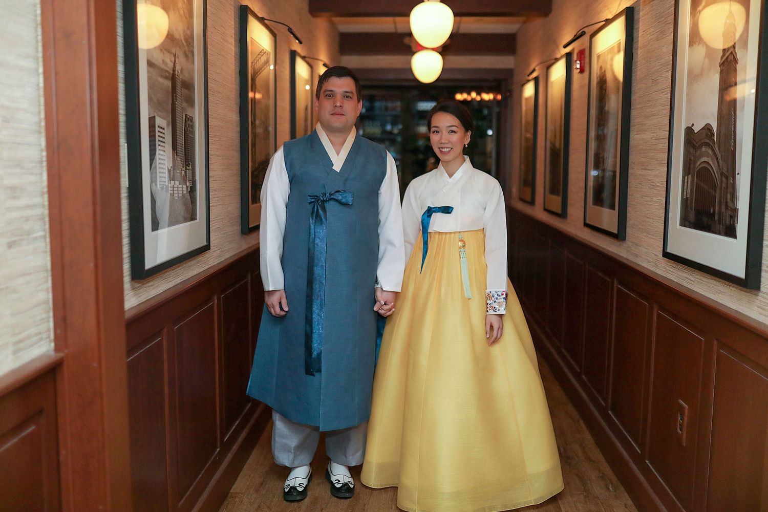 A couple walks down a vintage hallway decorated with framed artwork and illuminated by glowing wall sconces.