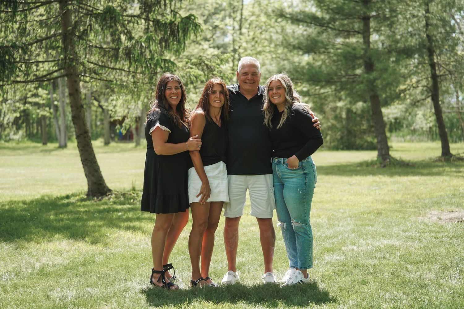 Four people stand together on a sunny lawn surrounded by evergreen trees in a casual outdoor setting.