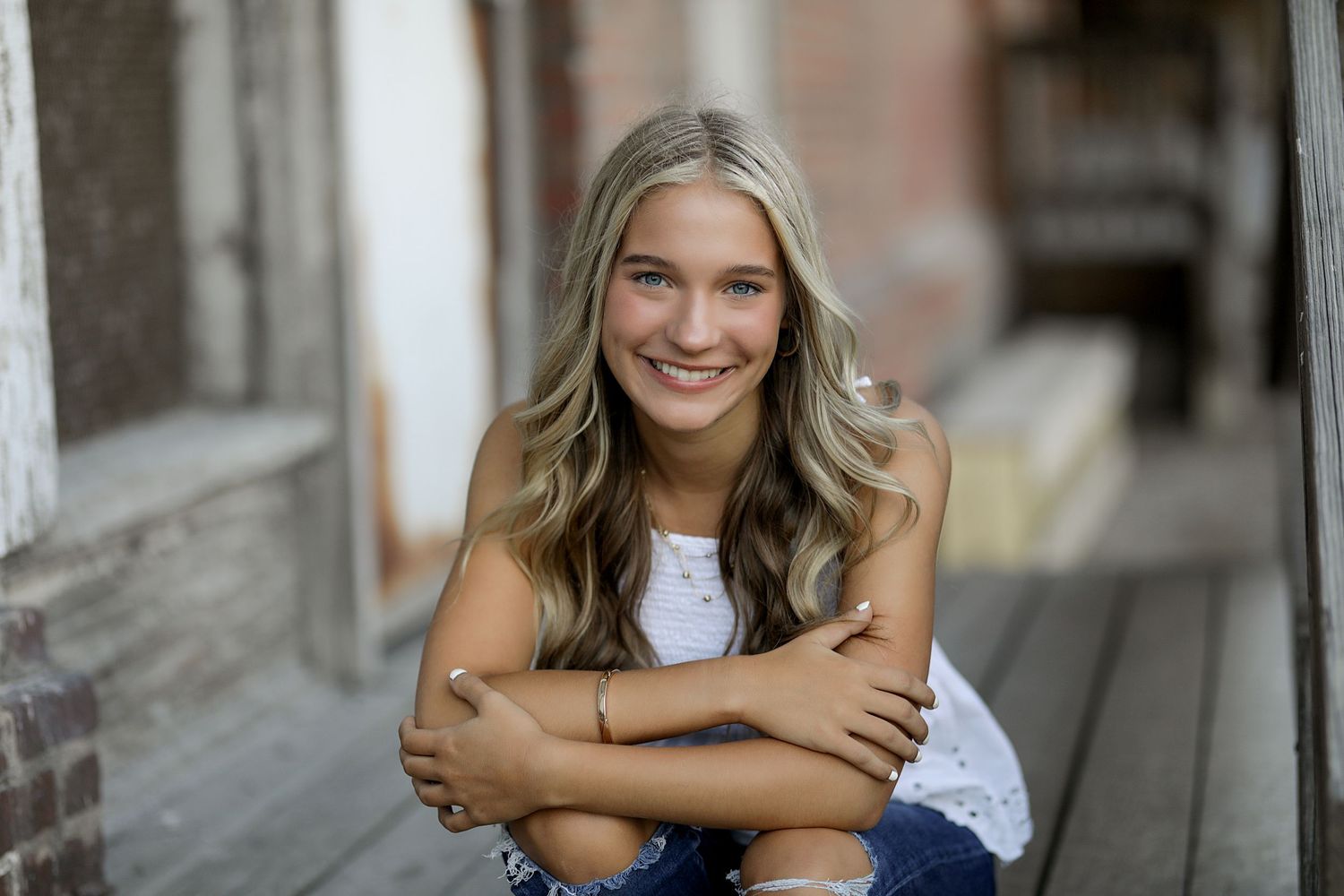 A person with wavy blonde hair wearing a white top and jeans sits outdoors smiling warmly with arms crossed.