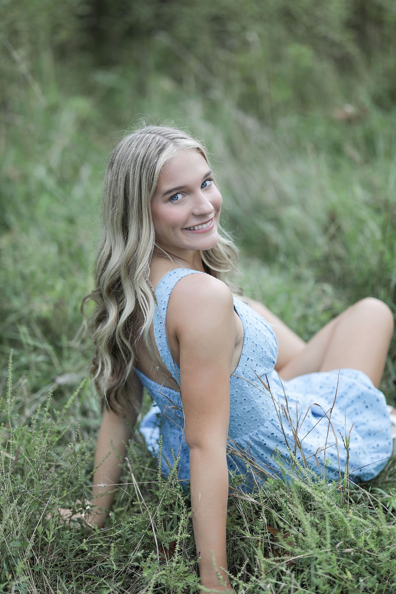 Person in light blue dress sits on grass with long wavy hair and natural outdoor backdrop in portrait setting.