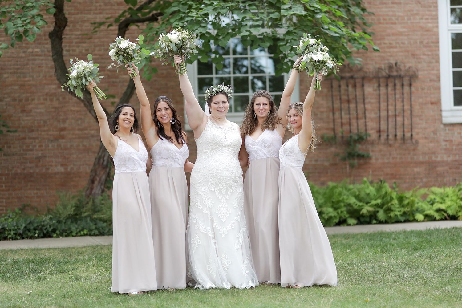 A bridal party wearing light beige gowns and holding white bouquets poses together on a lawn in front of a brick building.
