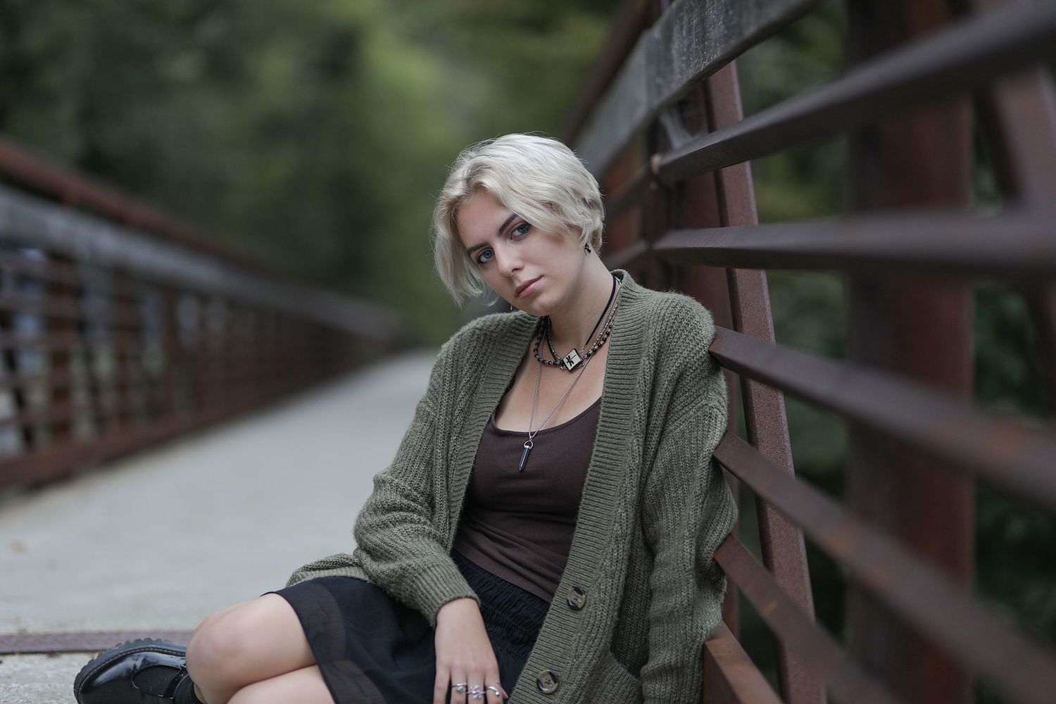 A person in an olive green cardigan and black dress sits thoughtfully on a bridge railing during an overcast day.