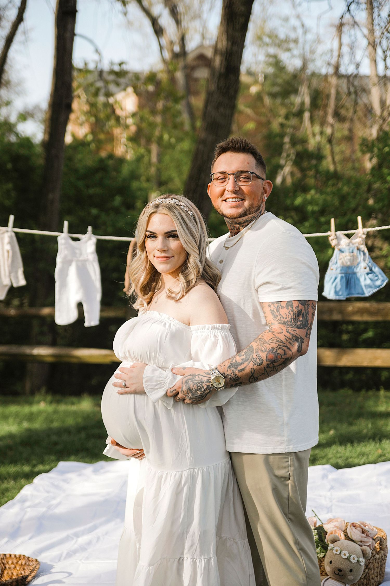 A couple poses in front of a clothesline with baby items hanging, during an outdoor maternity photoshoot in nature.