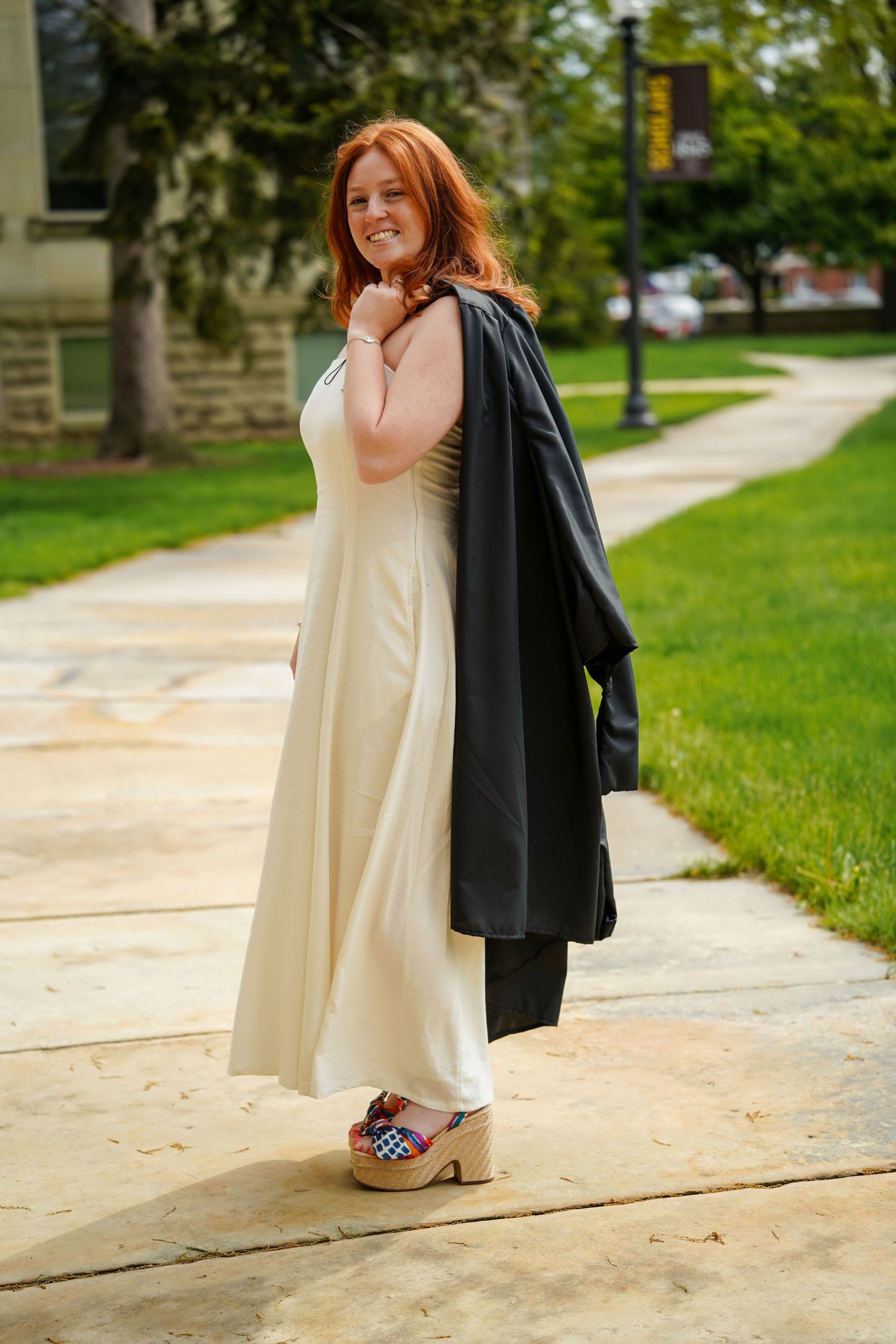A person in a cream-colored dress and platform shoes stands on a sidewalk holding a black graduation gown on campus.