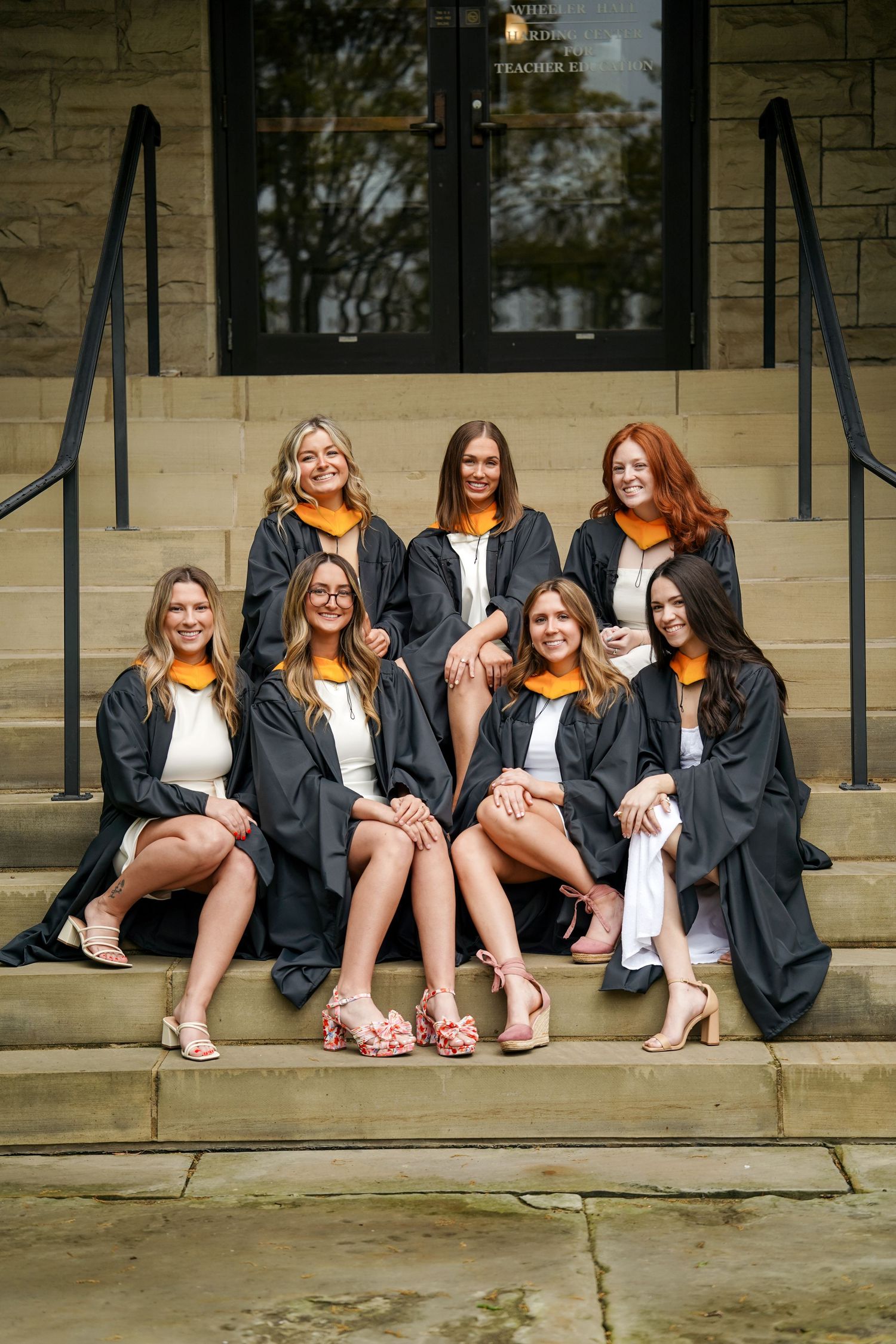 Group of graduates in black gowns showing their footwear while seated on church steps together.