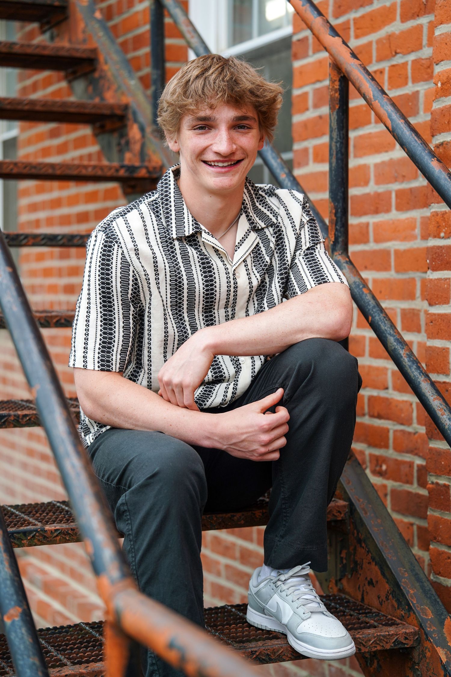 Person sitting on metal fire escape stairs wearing a striped button-down shirt and dark pants against brick building.