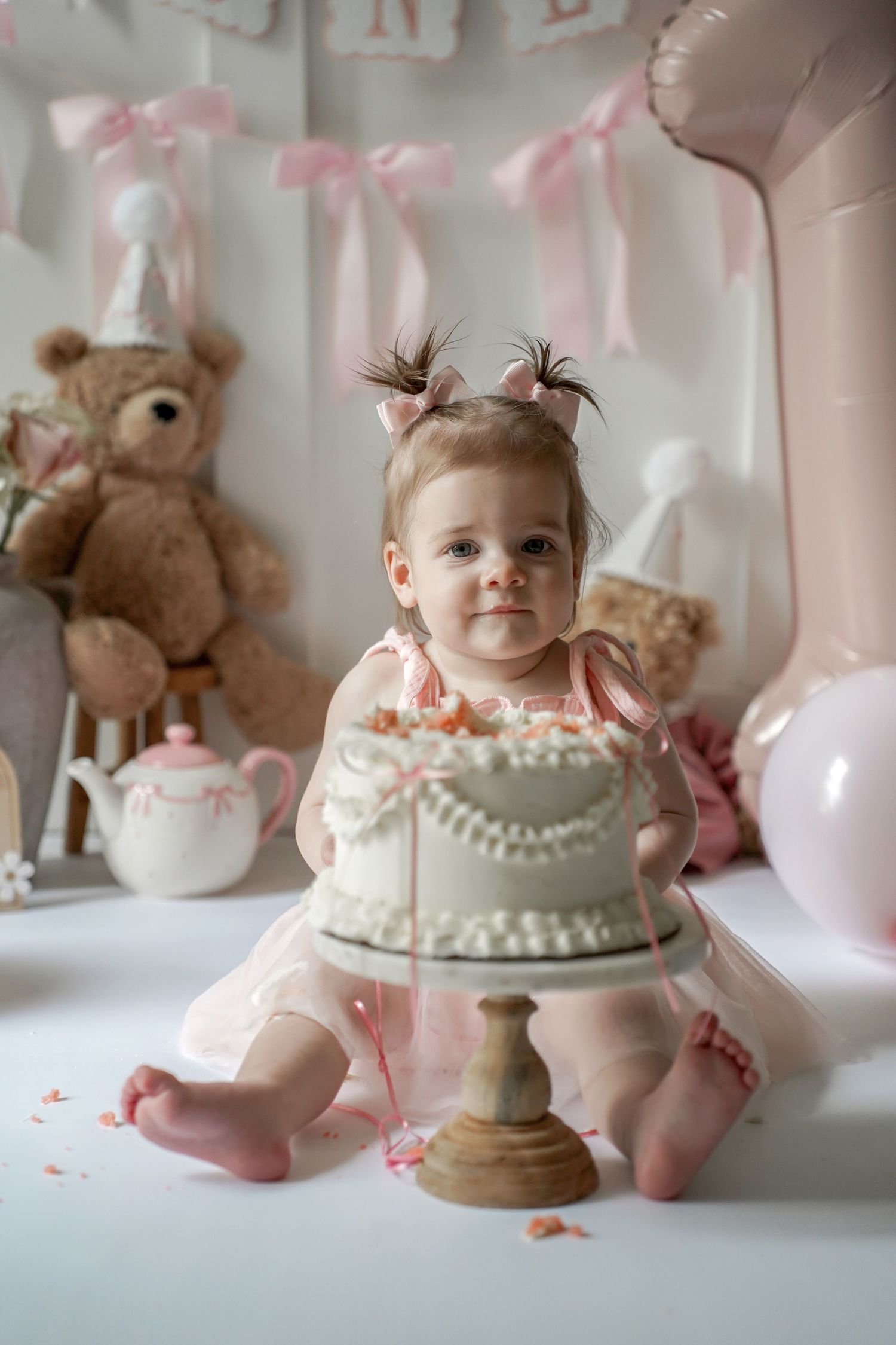 Pink and white decorated smash cake photo session setup with teddy bear and balloons in soft pastel colors.