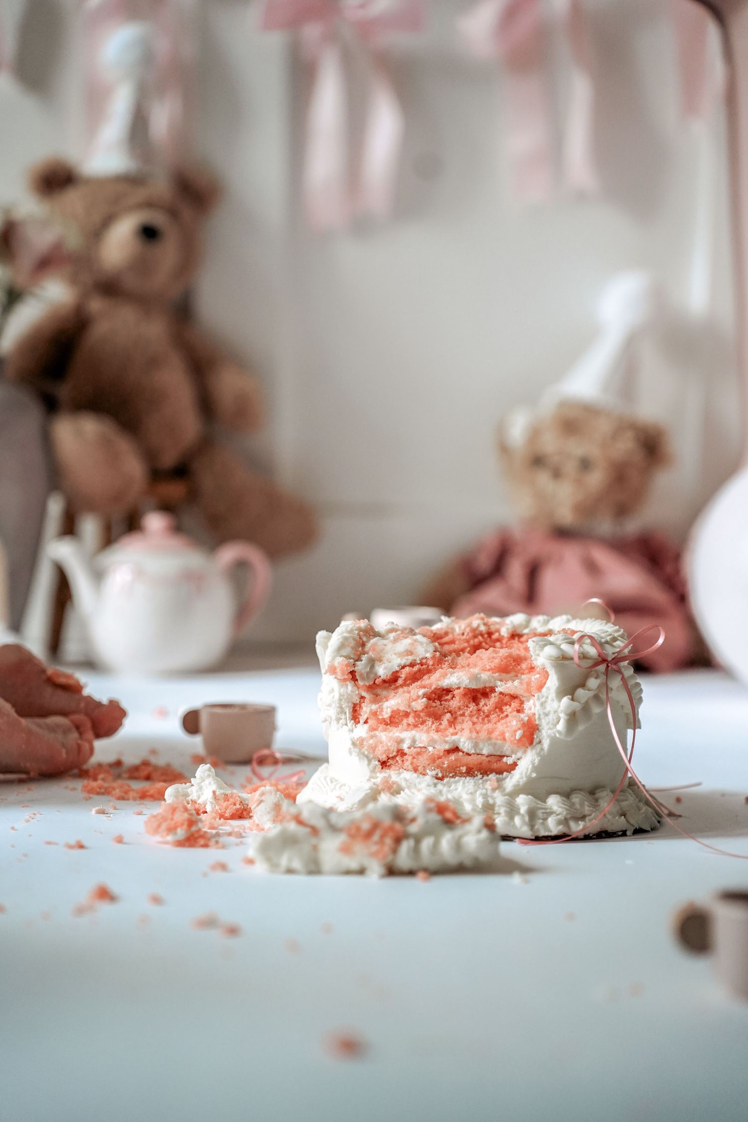 A pink cake with white frosting sits partially eaten on a table with teddy bears and tea set in soft focus background.