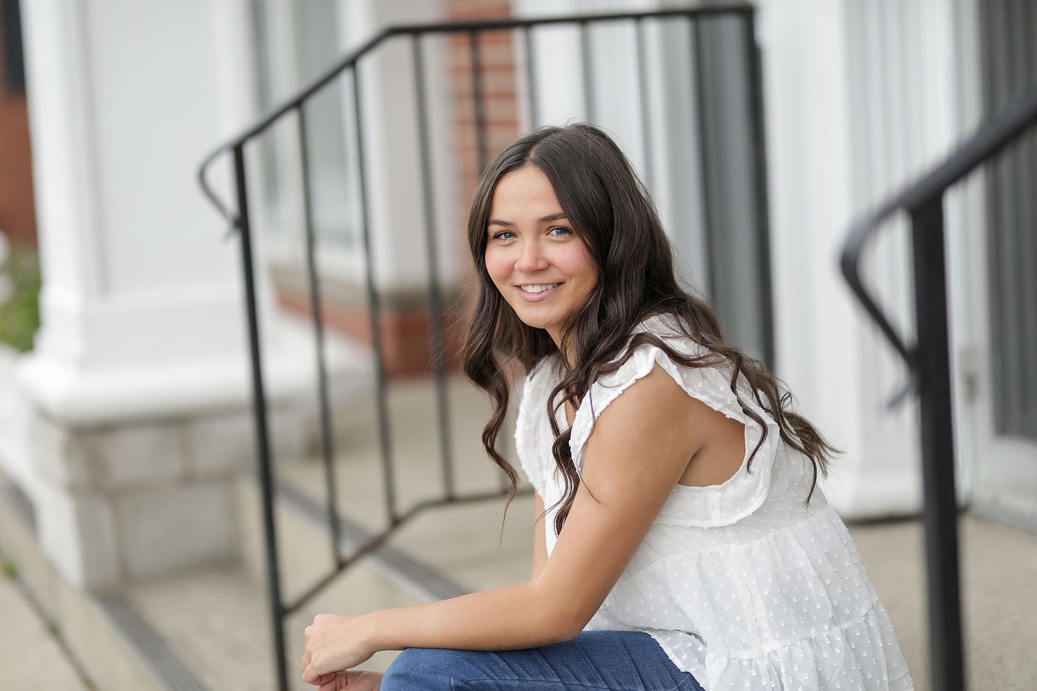 Young woman in white ruffled blouse and jeans sits casually on front steps, smiling warmly at camera.