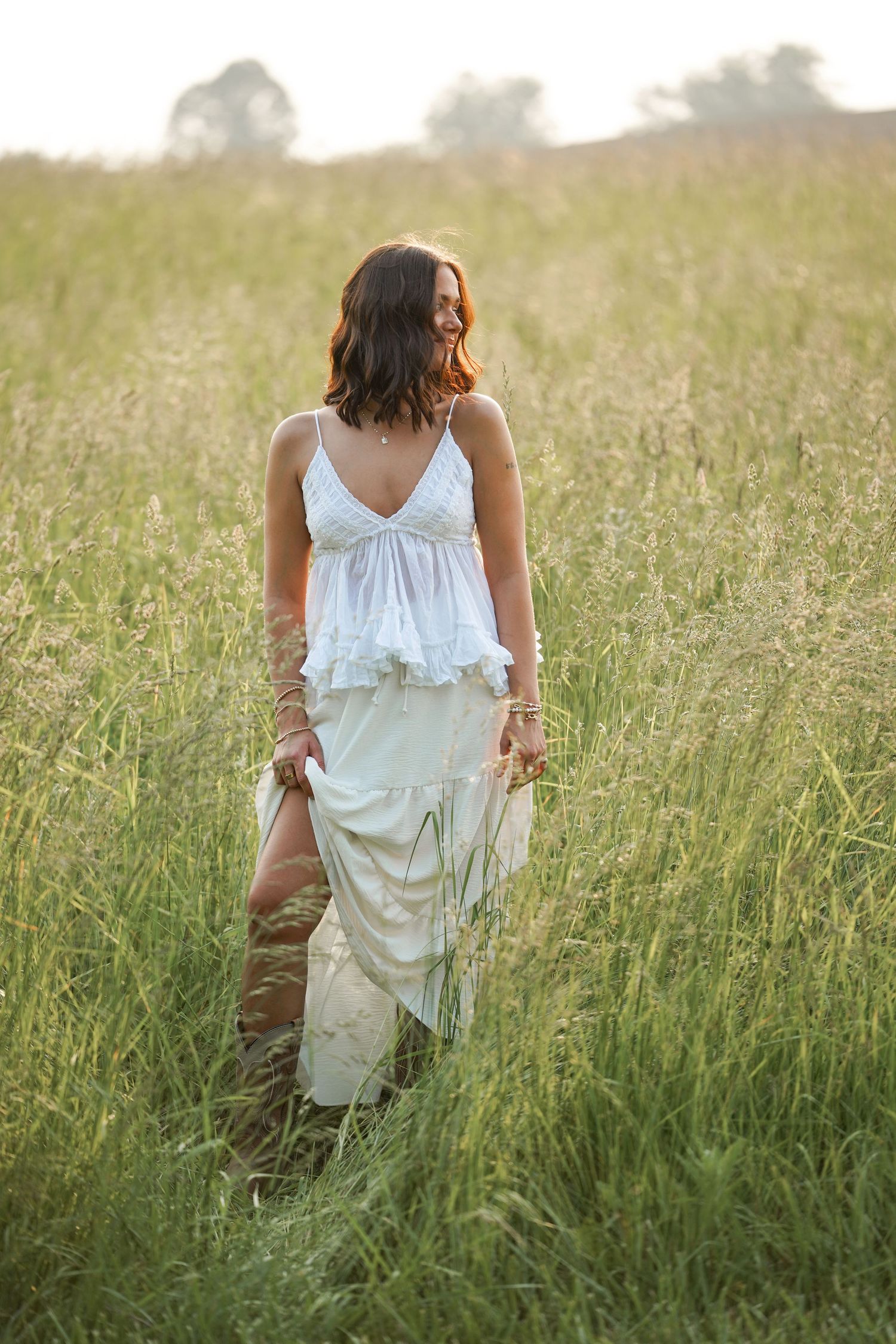 A white sundress-clad figure walks through tall golden grass in a sunlit meadow at dusk.